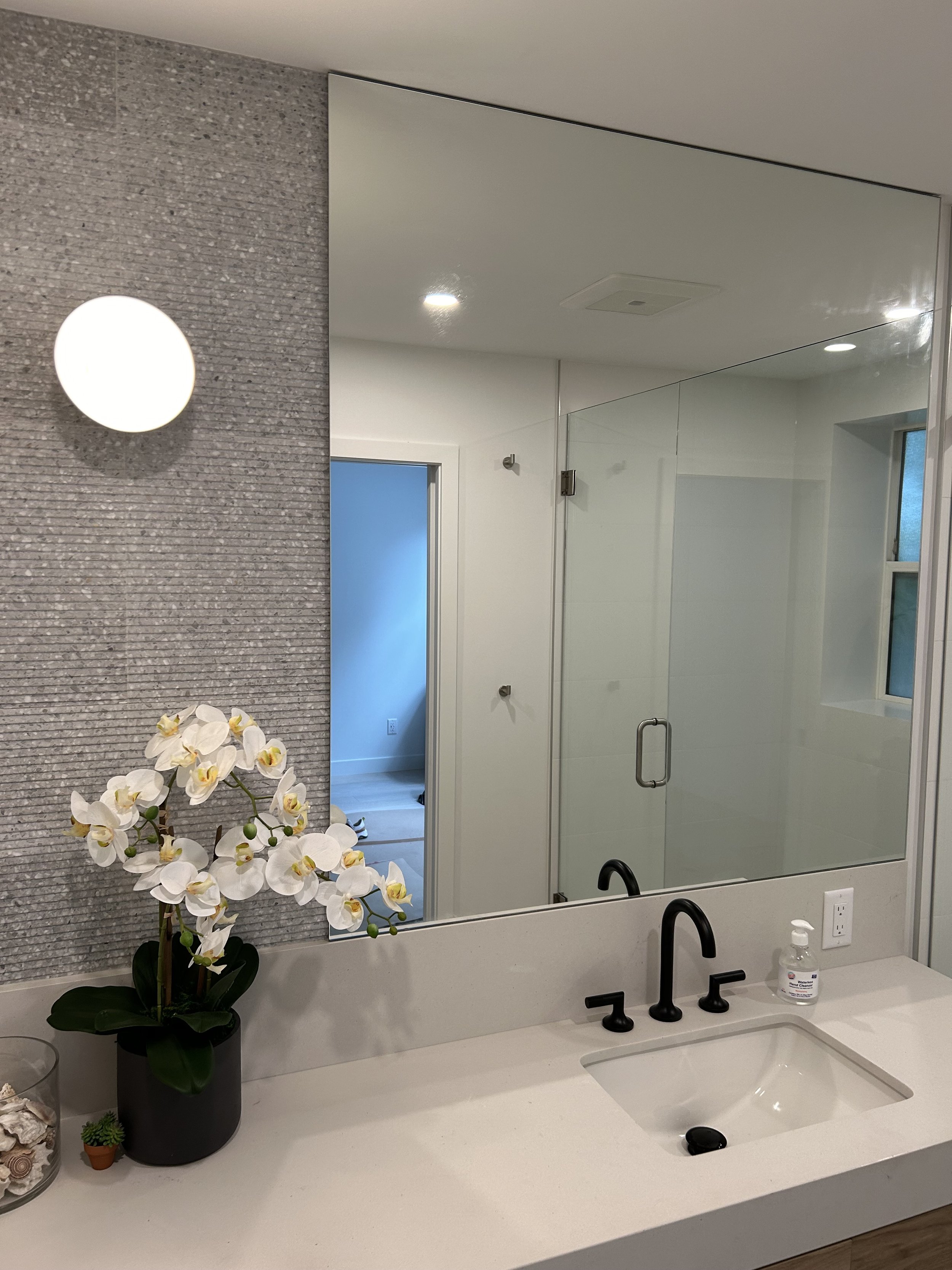 Bathroom with a large mirror above a white countertop, black faucet, and a potted orchid flower. The wall behind the mirror has textured gray tiles. To the right, there is a glass shower enclosure and a window. A bottle of hand soap is on the countertop.