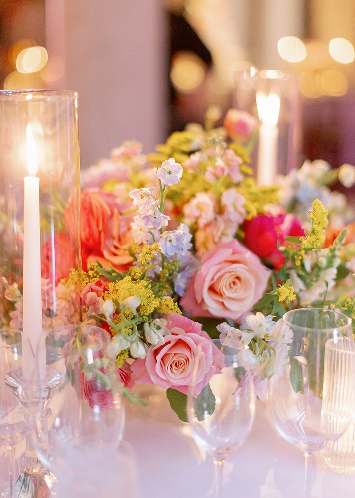 Elegant floral centerpiece with pink roses, white and yellow flowers, surrounded by candles with flickering flames on a table for a celebration or wedding.