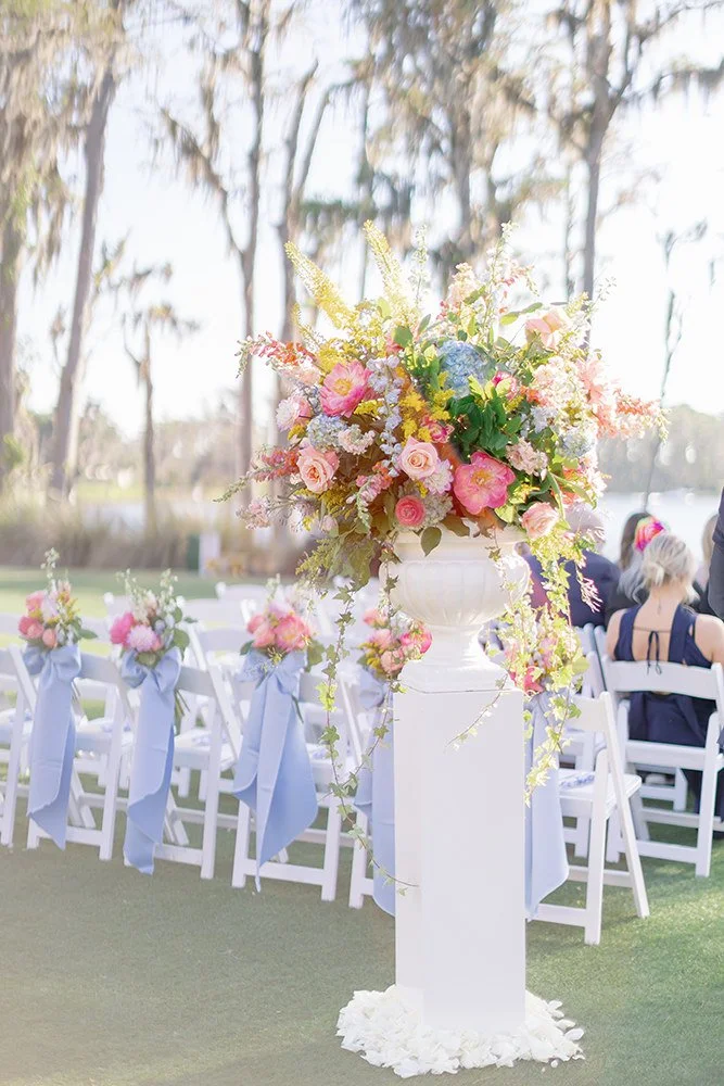 A large flower arrangement with pink roses, blue, yellow, and purple flowers, and green foliage in a white vase on a white pedestal at an outdoor wedding or event.