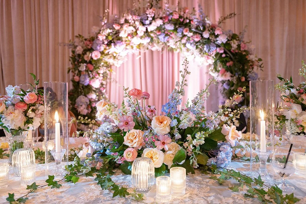 Elegant wedding table decorated with pastel-colored flowers, candles in glass holders, and a floral arch in the background.