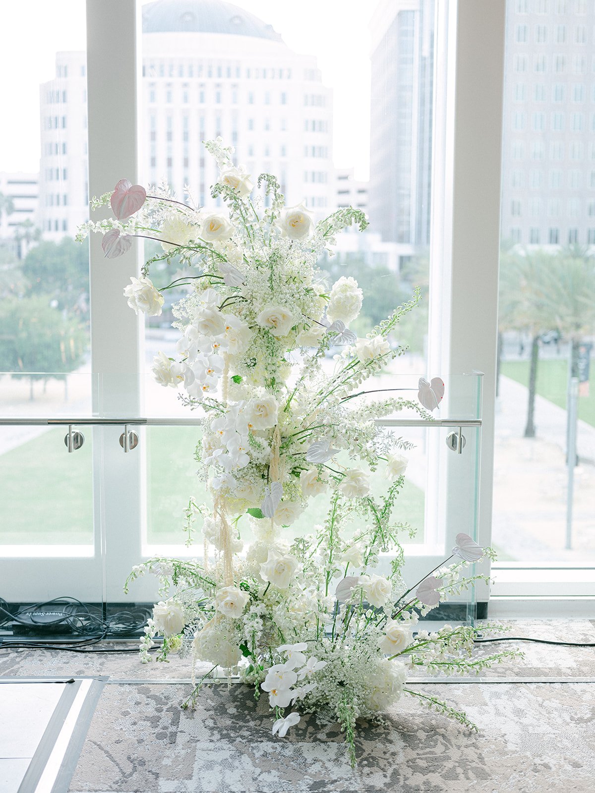 White floral arrangement with orchids, roses, and greenery in front of large windows with city buildings in background.