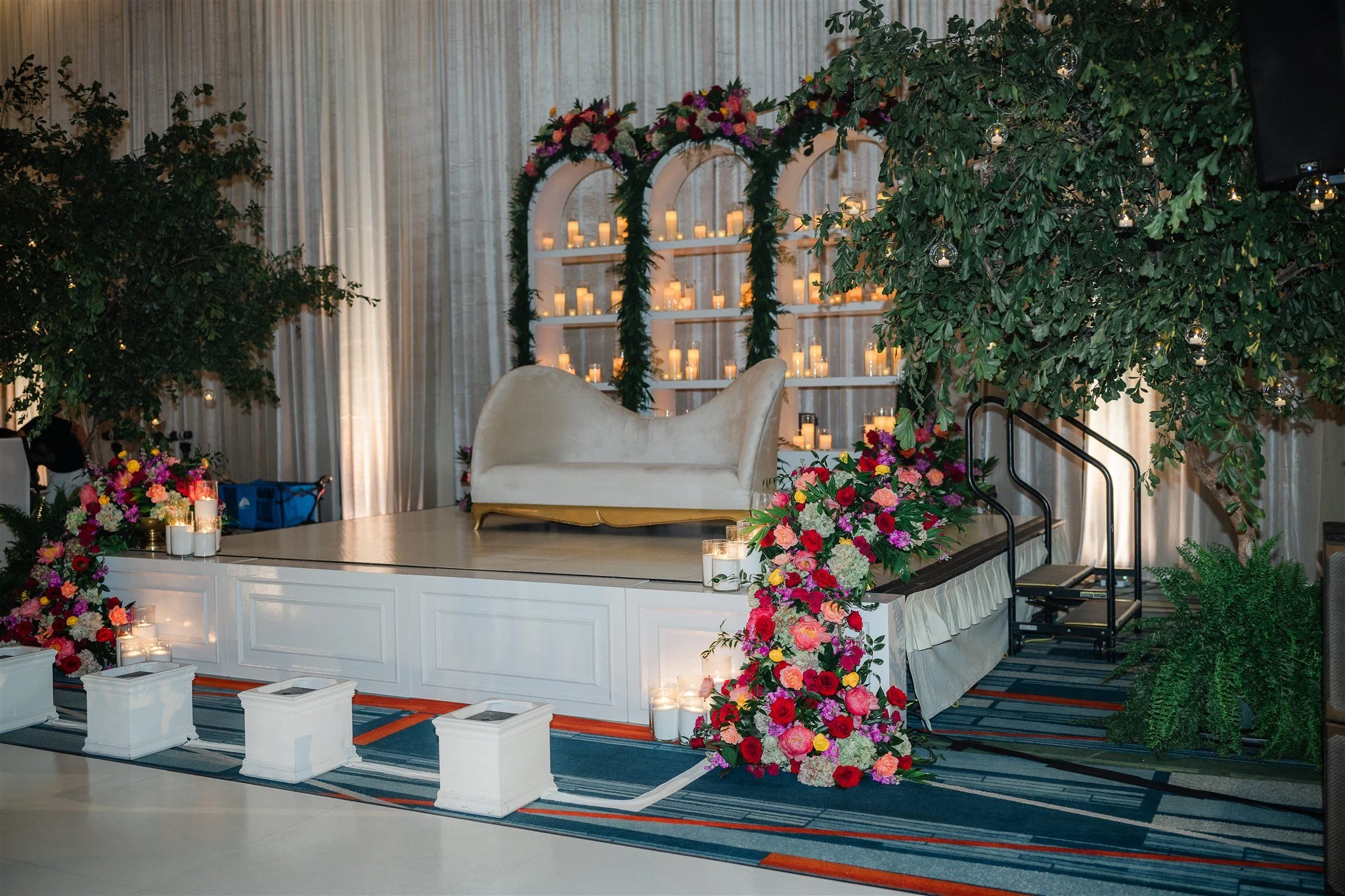 A stage decorated with numerous candles, colorful flowers, and green foliage, with a white sofa in the center, set up for a wedding or celebration.