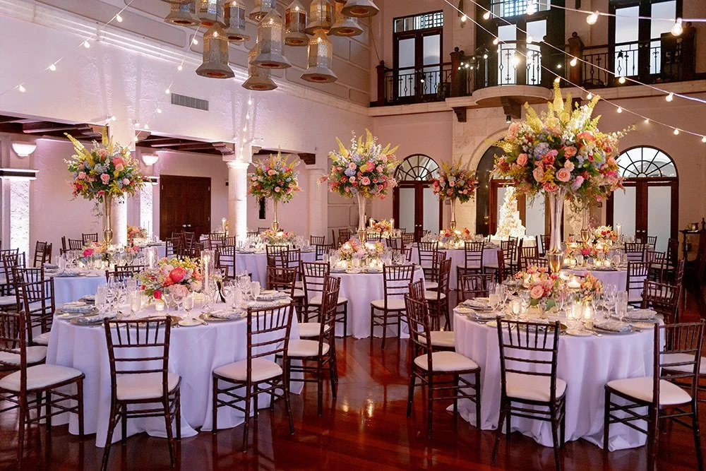 An elegant banquet hall decorated for a wedding or event, featuring round tables with white tablecloths, floral centerpieces in tall vases, and surrounded by wooden chairs. The room has high ceilings, large windows, and string lights hanging from the