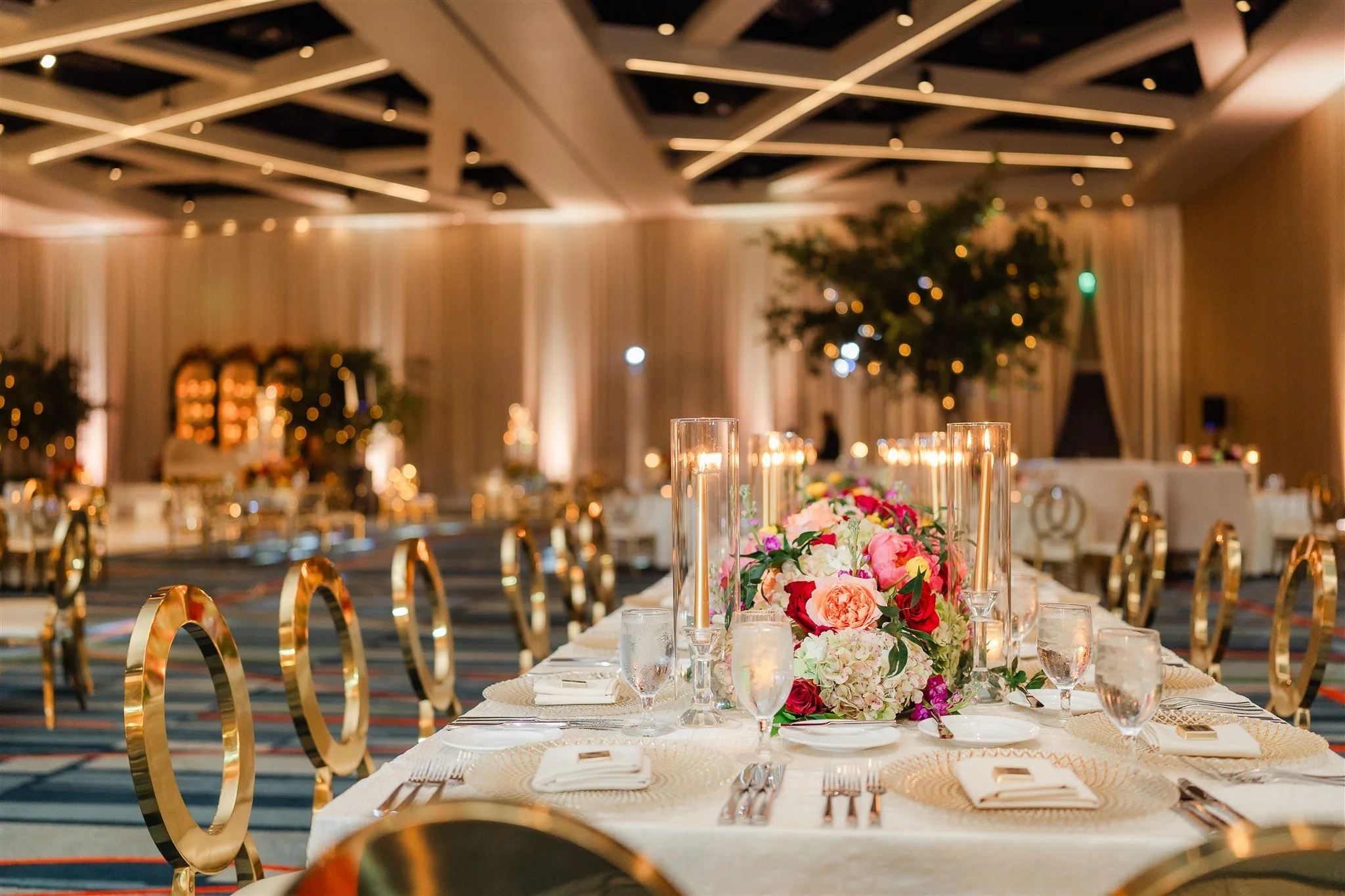 Elegant banquet hall with long table set with candles, glassware, and a floral centerpiece. The room has warm lighting, high ceilings with modern lighting design, and draped curtains in the background.