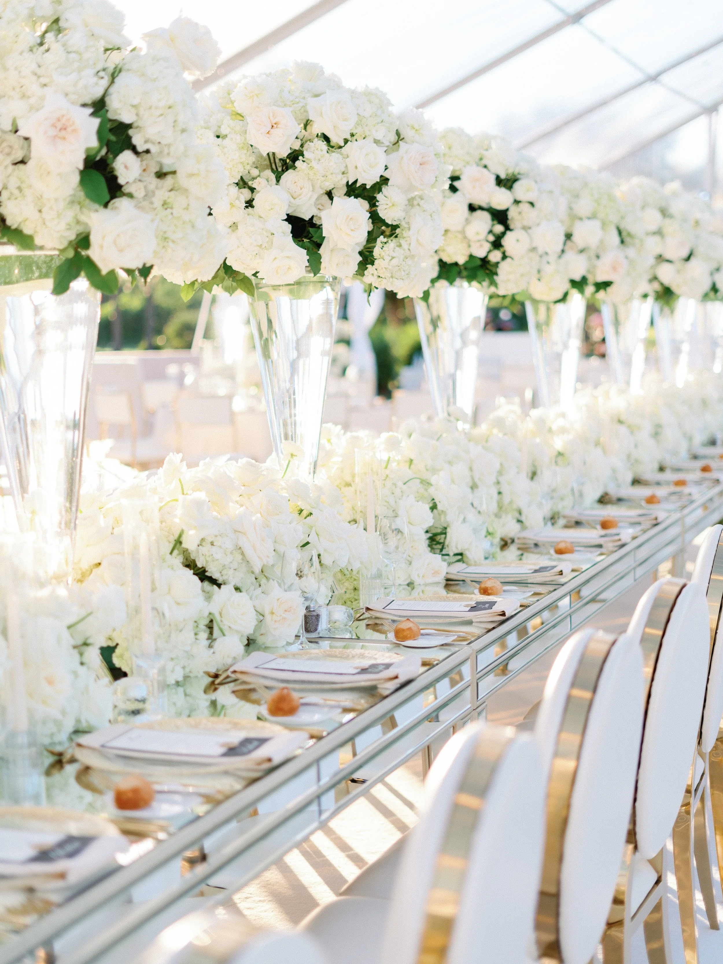 Elegant outdoor wedding reception table decorated with tall white flower arrangements in glass vases, set with white plates, napkins, and bread rolls, under a white tent with natural light.