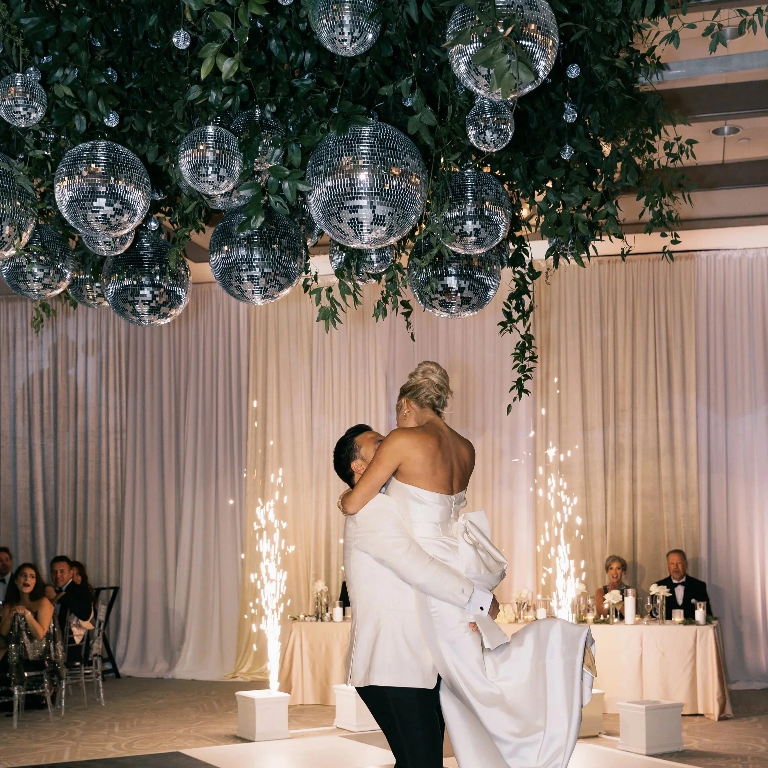 A couple is dancing at a wedding reception with disco balls hanging from the ceiling. Guests are seated at tables in the background, and elegant drapery decorates the space.
