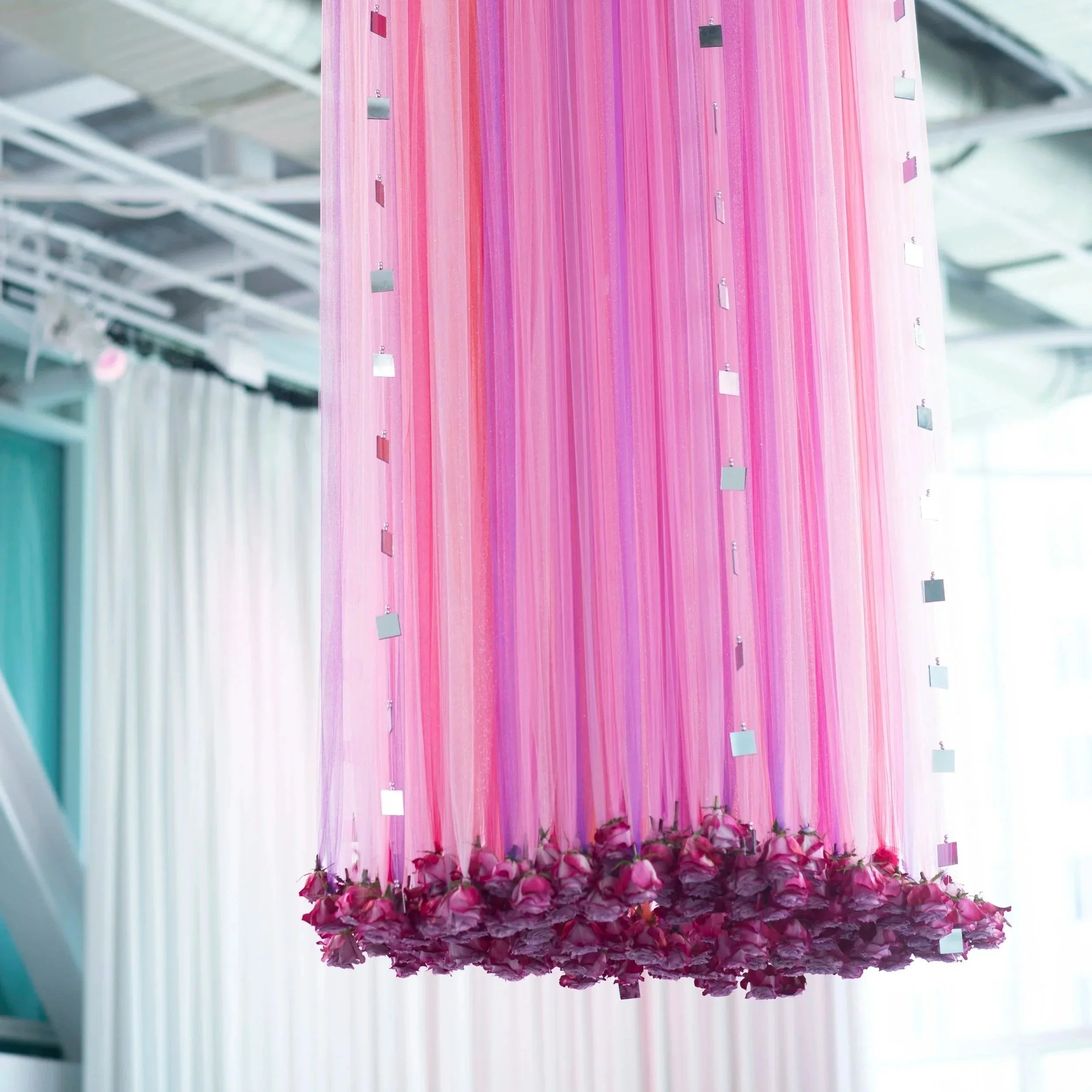 A colorful hanging installation with vertical pink and purple stripes, decorated with small reflective squares and surrounded at the bottom by pink flowers.