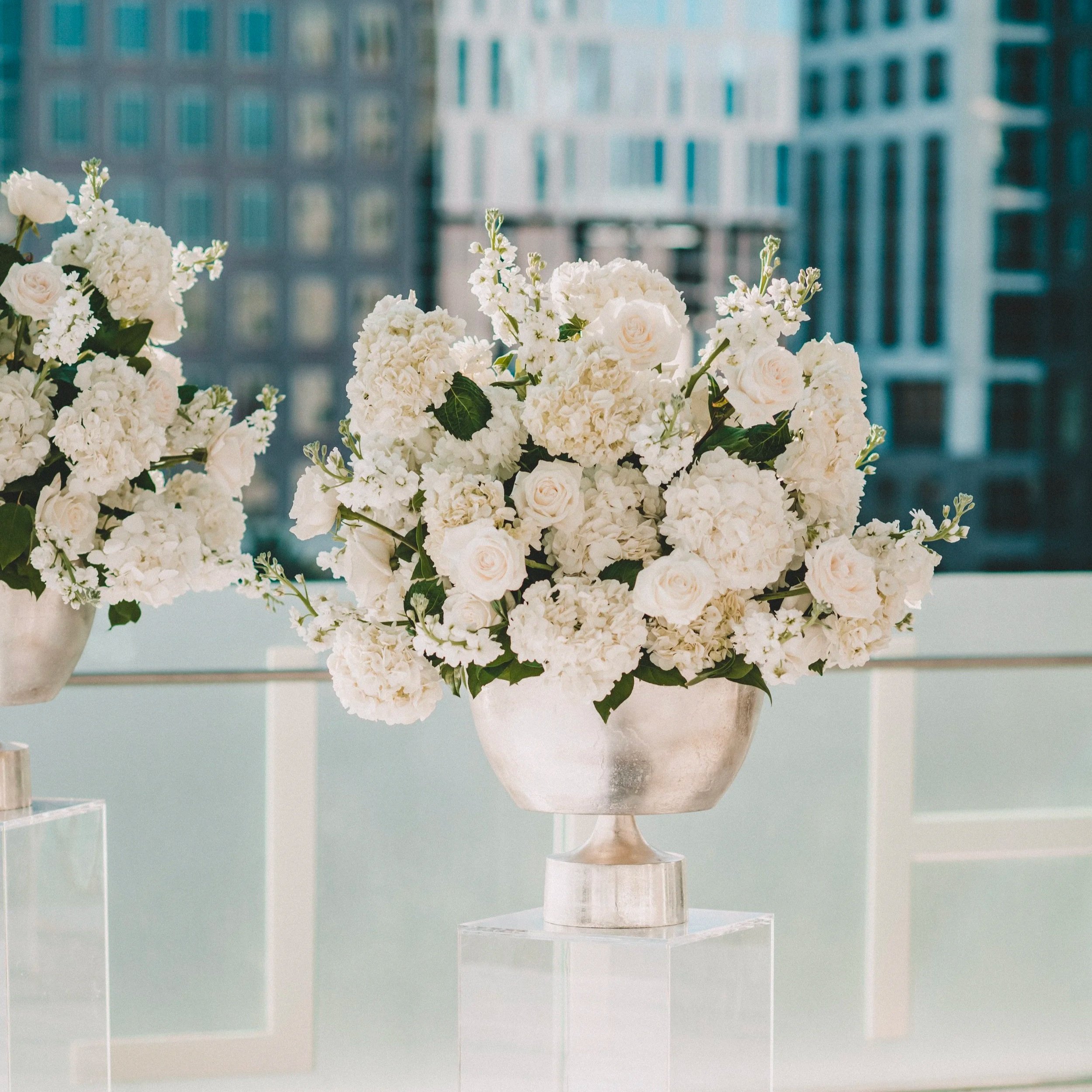 A floral arrangement with white flowers, including roses and hydrangeas, set in a decorative vase on a table with a cityscape blurred in the background.