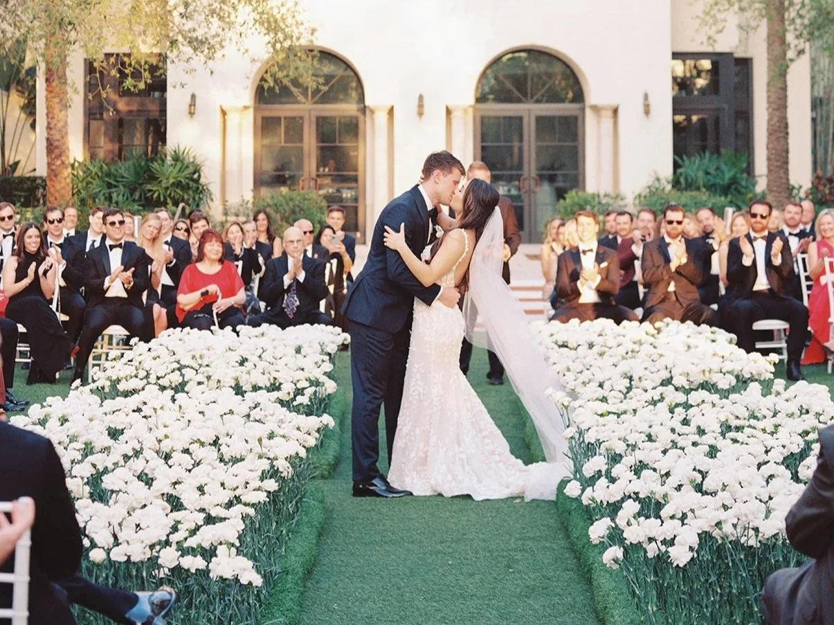 A couple kisses during their wedding ceremony outdoors, surrounded by guests and white flowers.