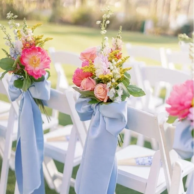 White folding chairs decorated with pink and white floral arrangements and light blue ribbons, set up outdoors for an event.