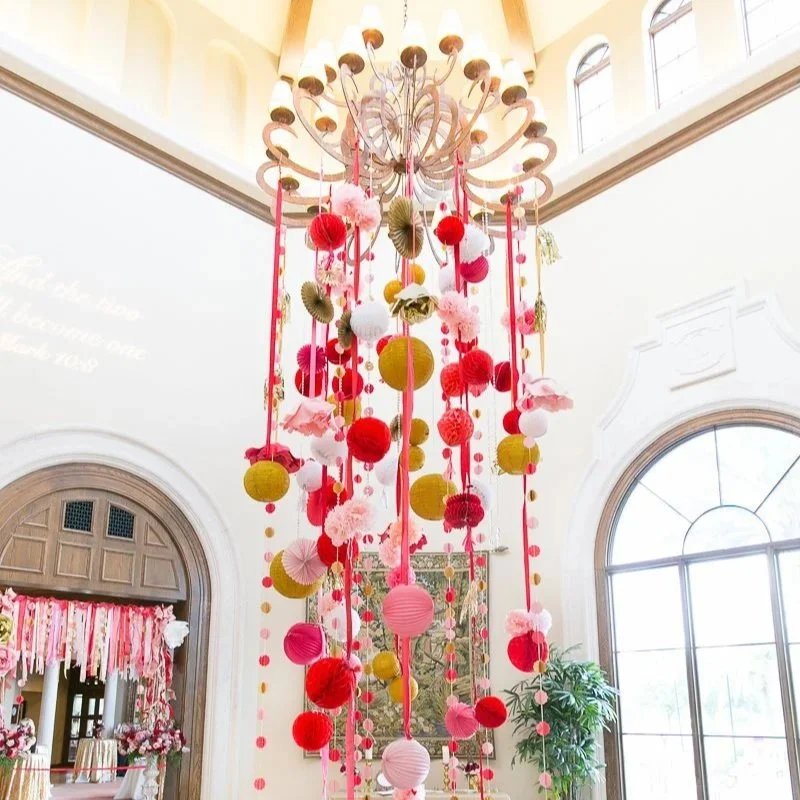 Colorful hanging paper decorations and pom-poms in pink, red, and gold, suspended from a chandelier in a bright, elegant room with large windows and white walls.