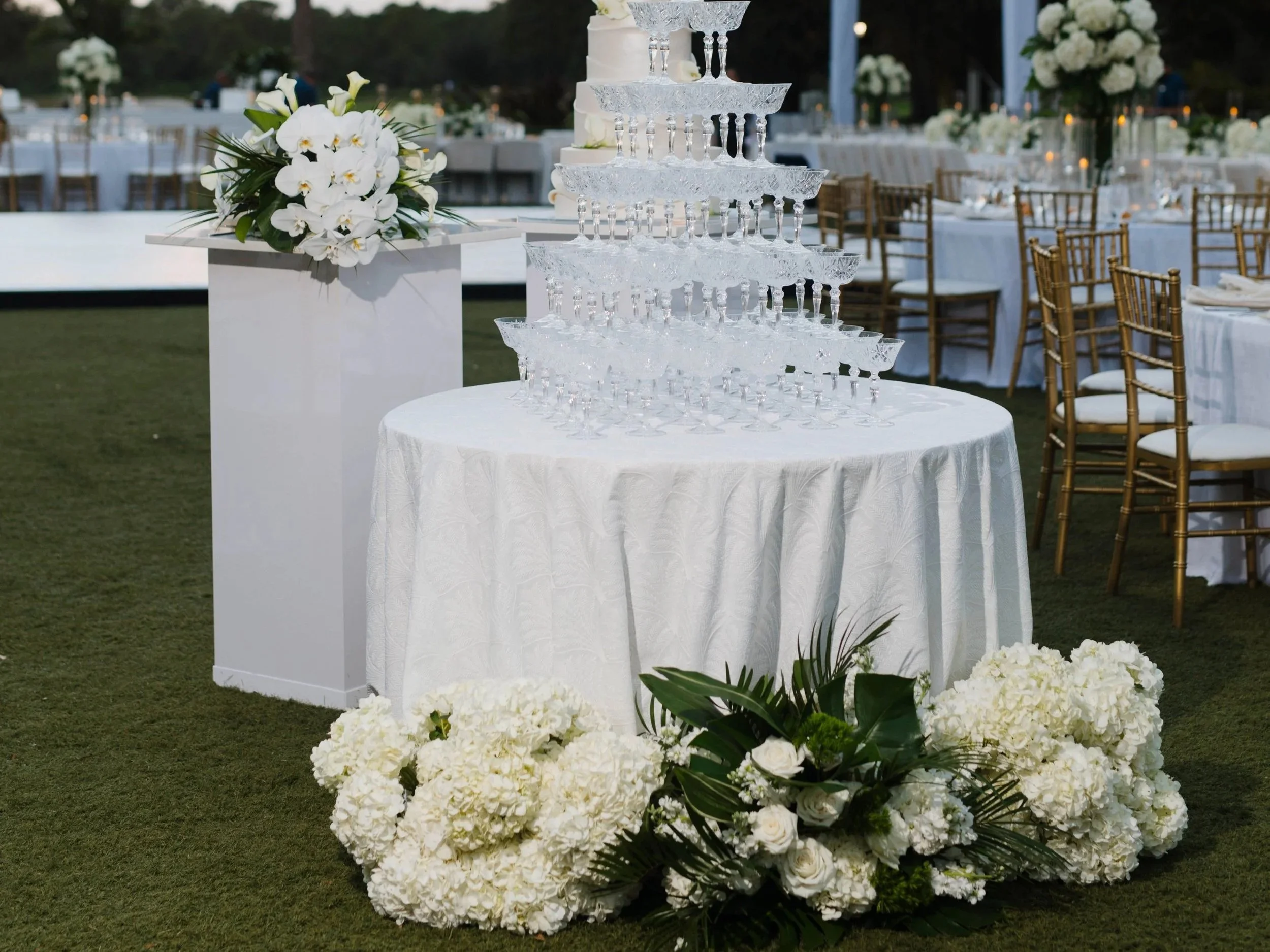 Elegant outdoor wedding reception setup with a tiered champagne tower, floral arrangements with white flowers, and twinkling string lights overhead at sunset.