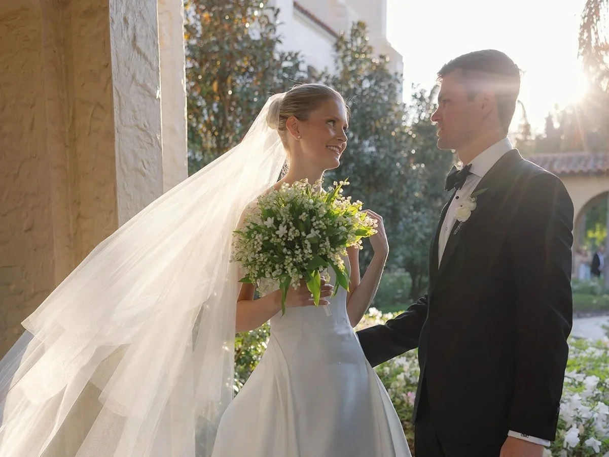 Bride and groom on wedding day outdoors, smiling at each other, with sunlight shining behind them, bride holding a bouquet of flowers.