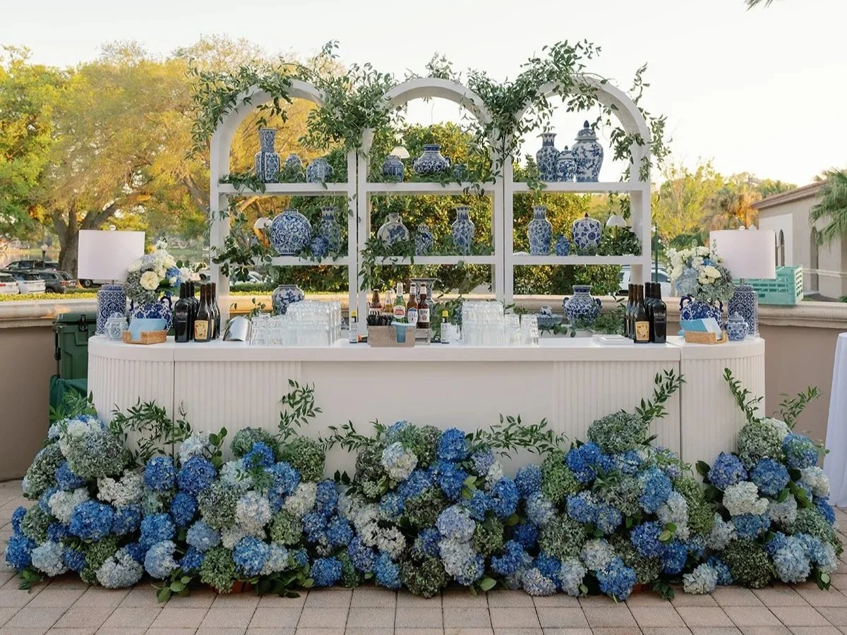 Wedding or event bar setup with blue and white floral arrangements, decorative blue and white vases, and assorted bottles and glasses on a white counter, with greenery and blue hydrangea flowers in front, outdoors with trees in the background.