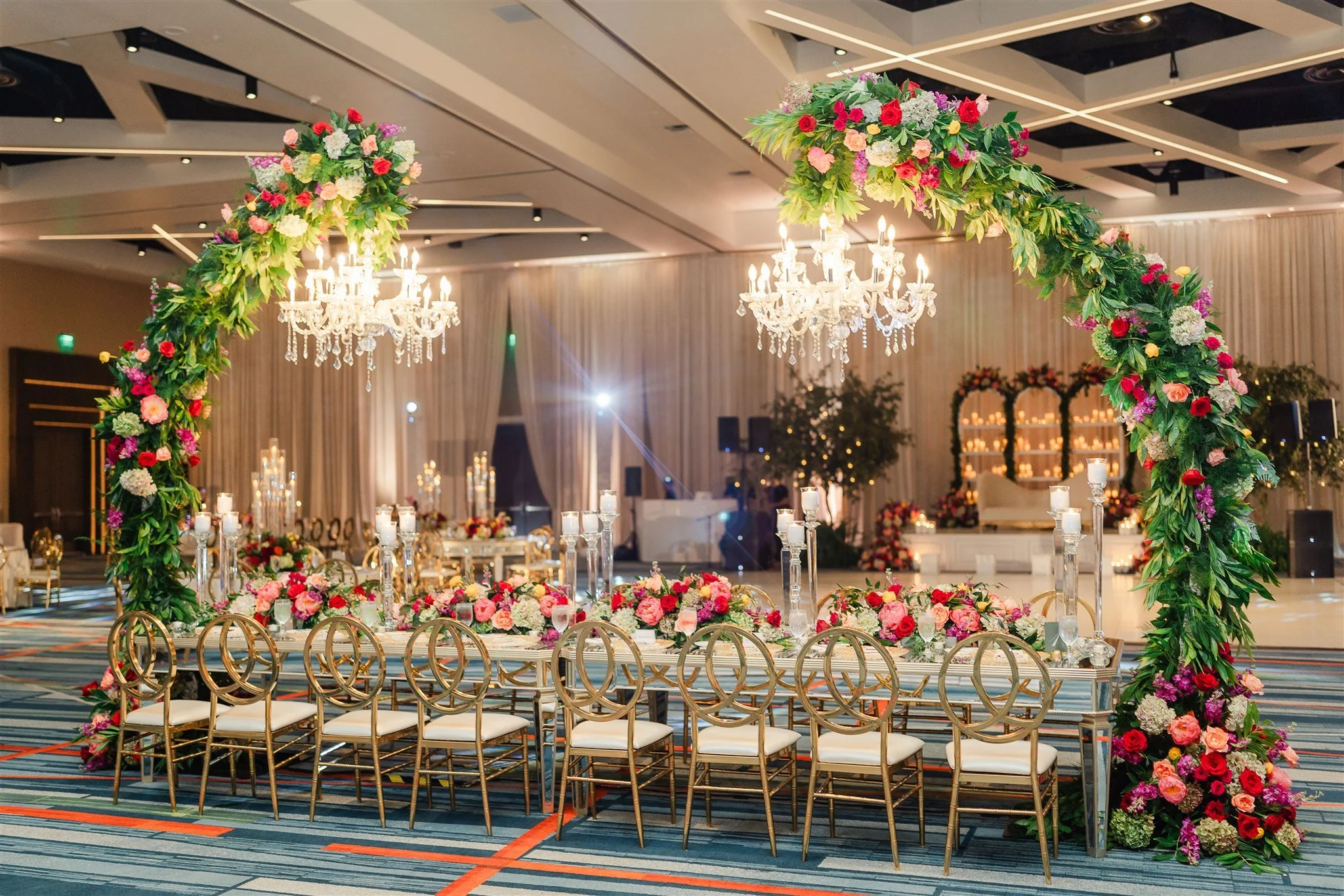 Elegant wedding reception setup with a large floral arch, crystal chandeliers, and decorated tables in a modern banquet hall.