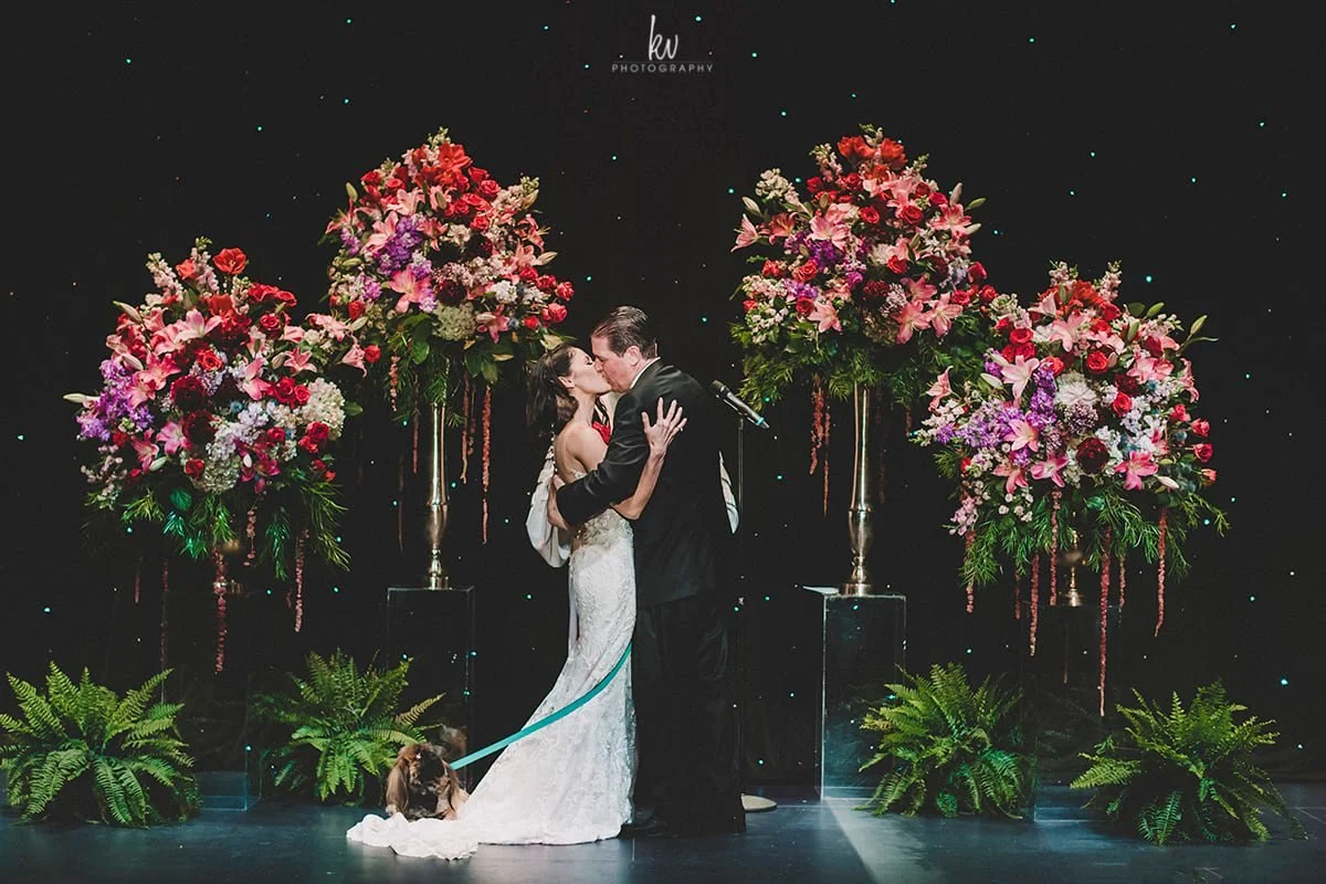 A bride and groom sharing a kiss on stage at their wedding, surrounded by four large floral arrangements with pink, red, and purple flowers, and green plants at their feet. A dog leash is held by the bride, with a dog lying on the floor.