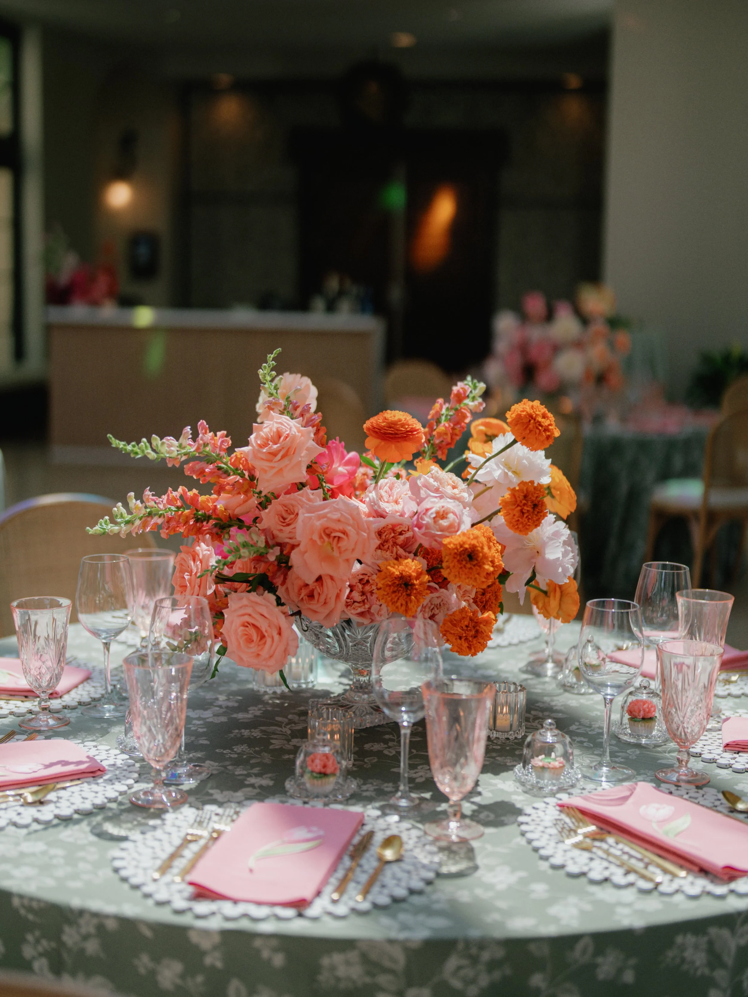 A centerpiece of pink, orange, and white flowers on a table set for a formal dinner.