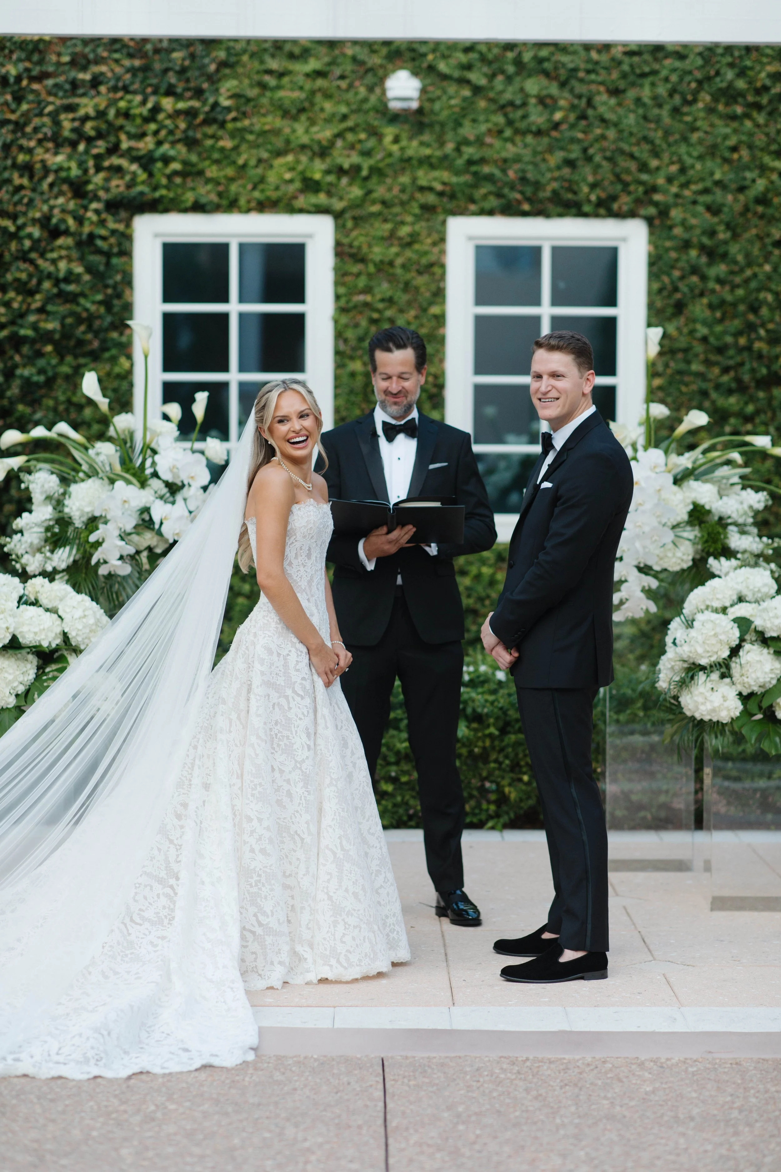 A bride and groom exchanging vows at a wedding ceremony outdoors, with a person officiating and a background of greenery and white flowers.