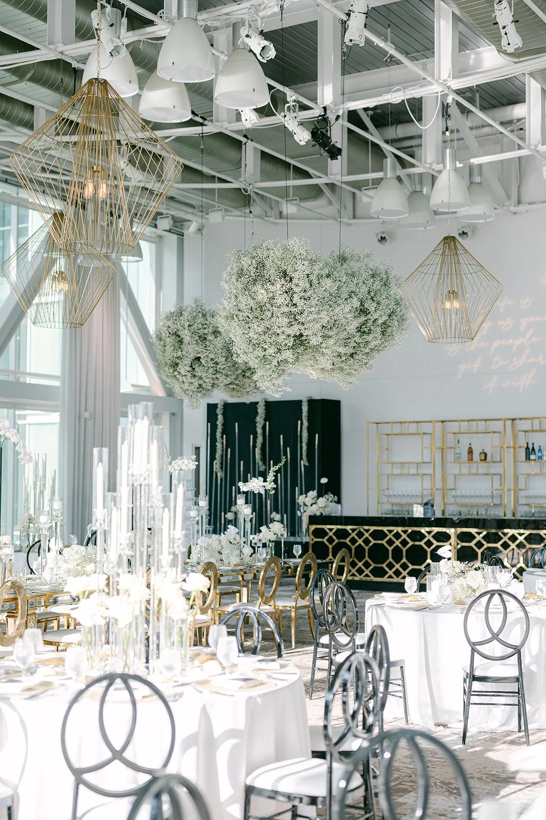 Elegant event space with white tables, floral centerpieces, modern chairs, hanging floral arrangements, large crystal chandeliers, and decorative lighting fixtures.