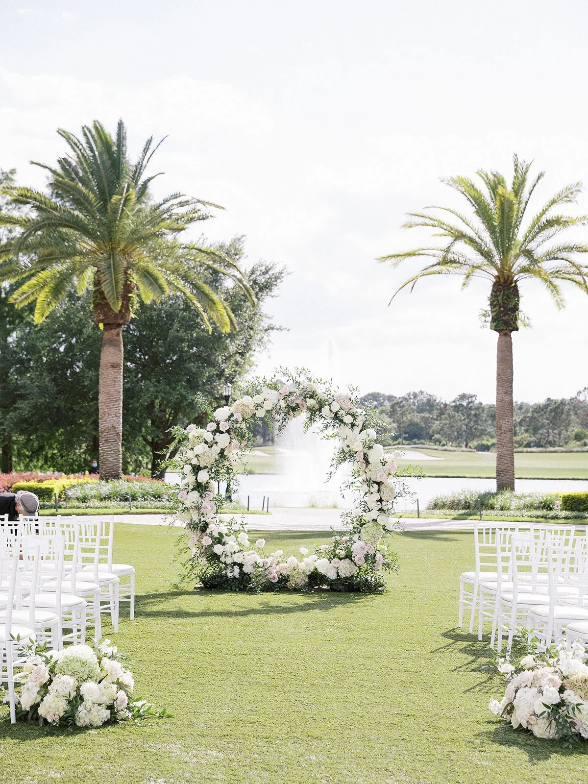 Outdoor wedding setup with white chairs, floral arch, palm trees, lake view, and fountain in the background.