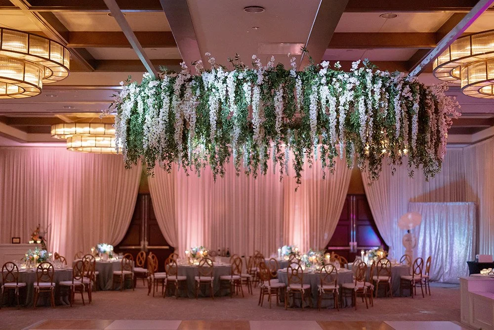 Elegant banquet hall decorated with hanging floral arrangement, draped curtains, and round tables with centerpieces, warm lighting, and subdued pastel color scheme.