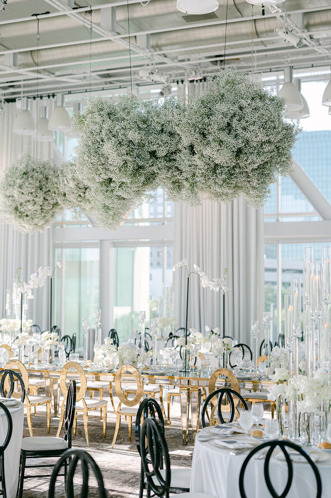 Elegant event space with hanging white floral arrangements, round tables covered with white tablecloths, black and gold chairs, and tall glass candle holders, in a bright venue with large windows and modern decor.