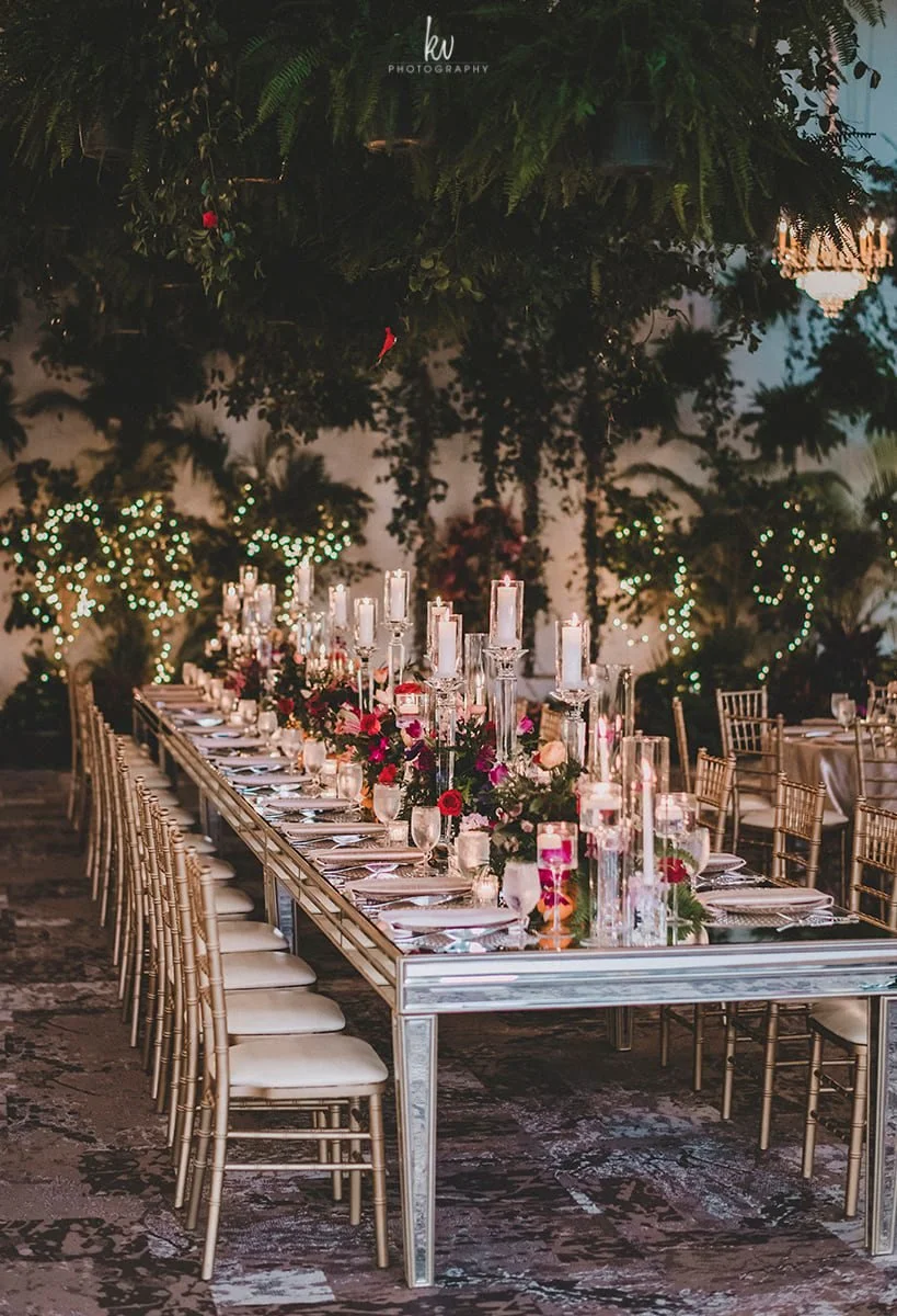 Elegant wedding reception table setup with candles and floral arrangements outdoors, surrounded by string lights and lush greenery.