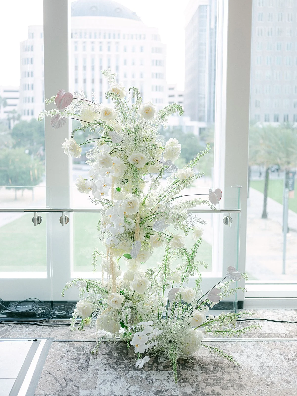 A large floral arrangement of white flowers and greenery on a stand in front of a large window with a city view.