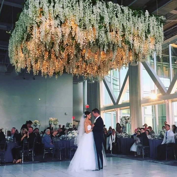 Bride and groom dancing at their wedding reception with guests seated at decorated tables in the background, large floral chandelier hanging above, and floor-to-ceiling windows allowing natural light.
