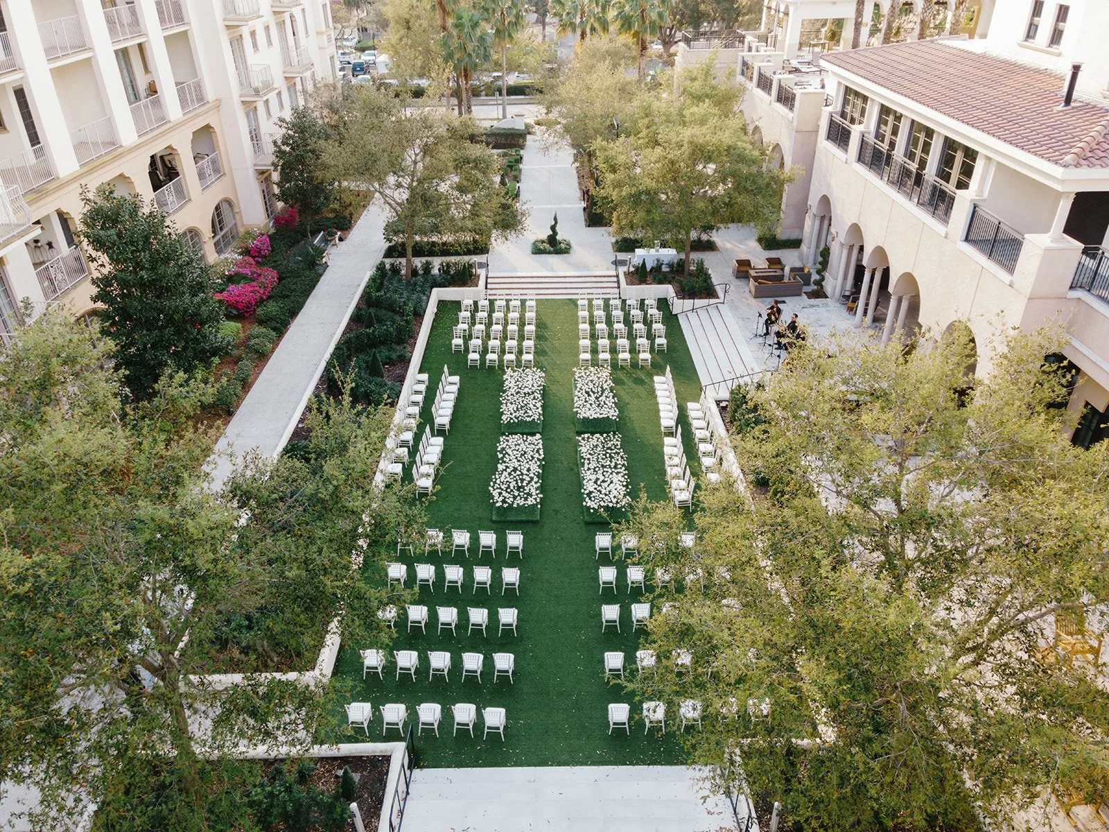 Aerial view of an outdoor wedding ceremony setup in a courtyard. White chairs are arranged on a grassy area with flower arrangements, surrounded by trees and multi-story buildings.