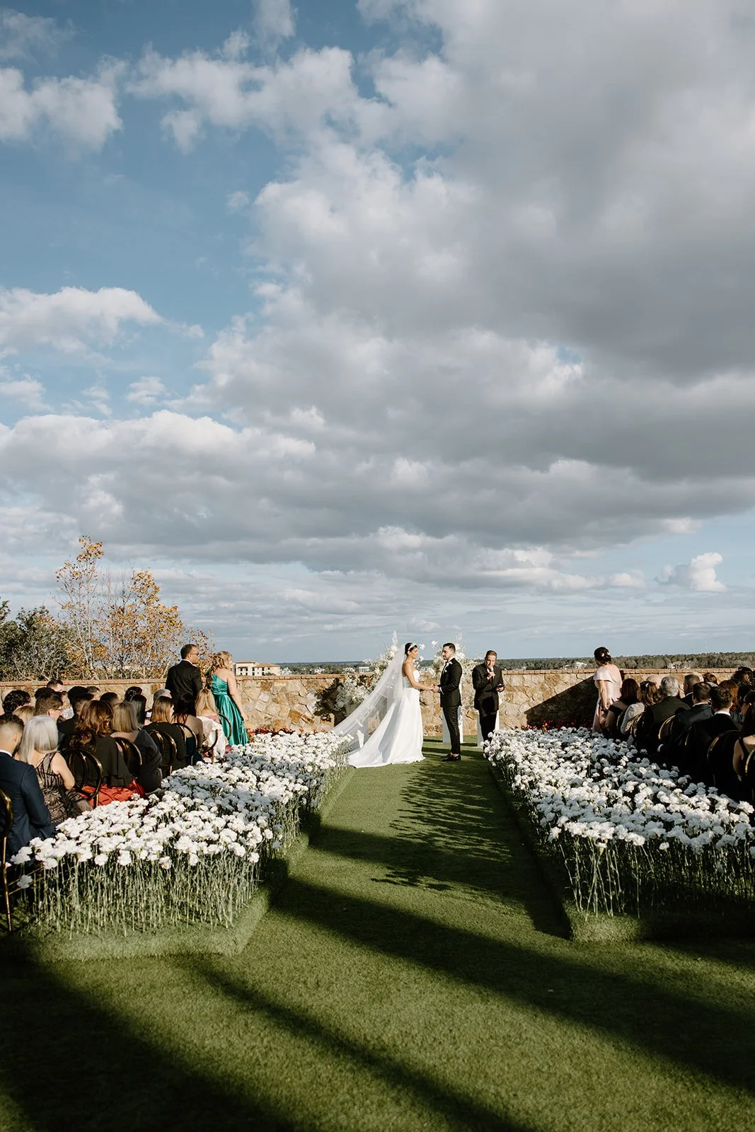 Outdoor wedding ceremony with a bride and groom exchanging vows on a grassy aisle decorated with white flowers, guests seated on either side, and a partly cloudy sky overhead.