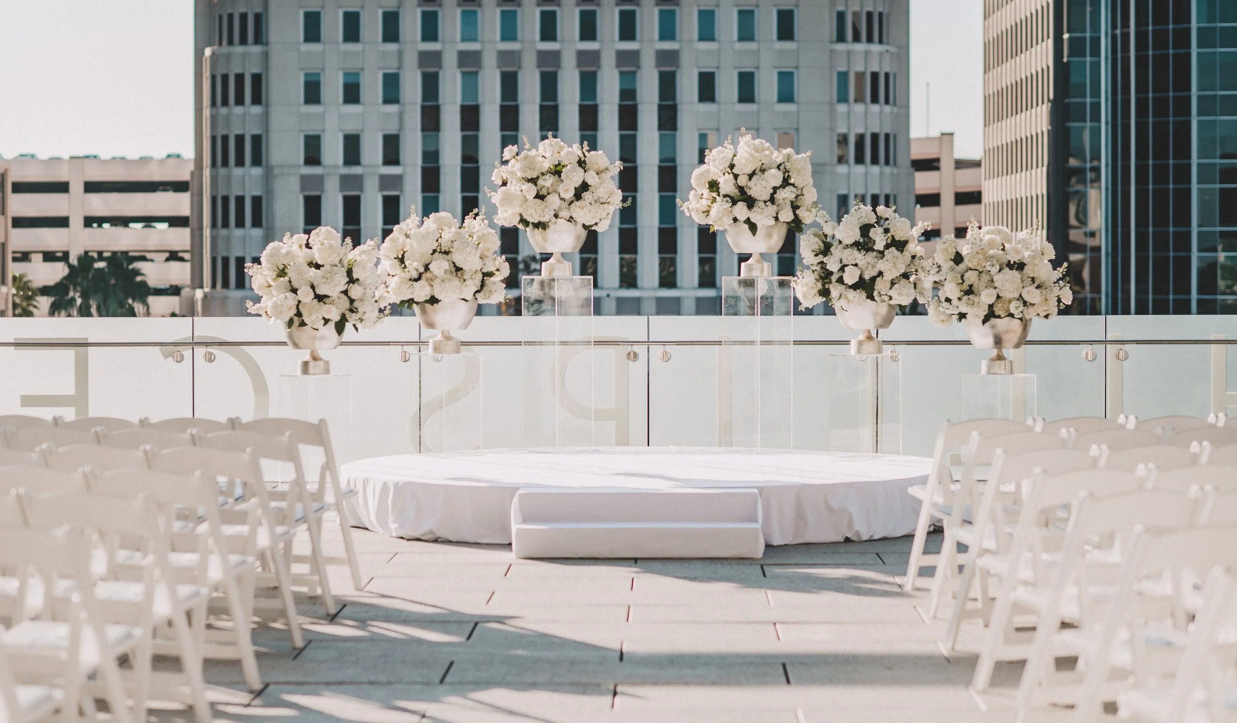 A round table with white chairs around it, decorated with five large floral arrangements in white vases on transparent pedestals, set on an outdoor terrace with a cityscape background.