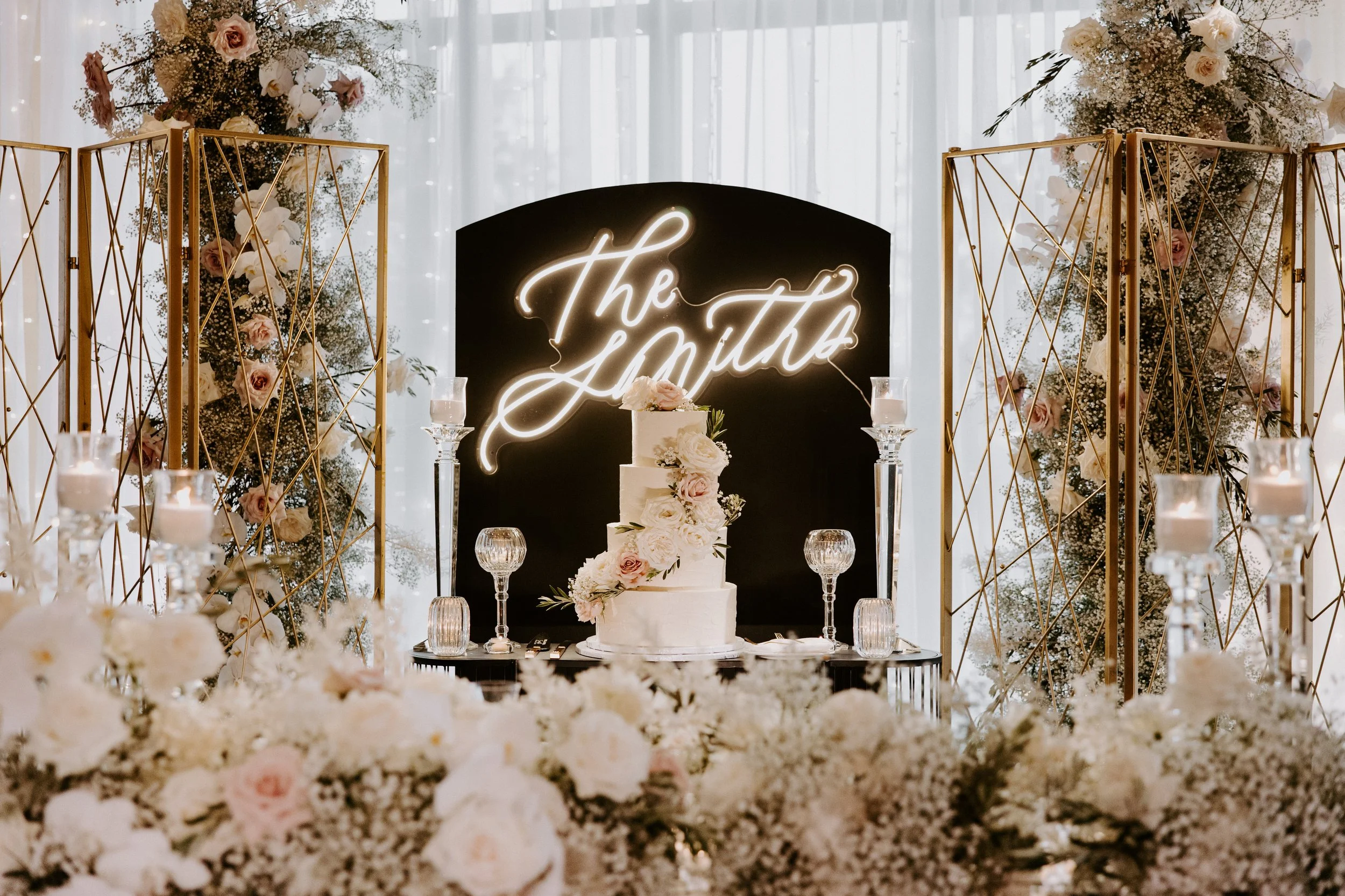 A wedding reception setup featuring a white three-tiered wedding cake decorated with flowers, surrounded by candles and glass holders, with floral arrangements and a neon sign that says 'The Smiths' in the background.