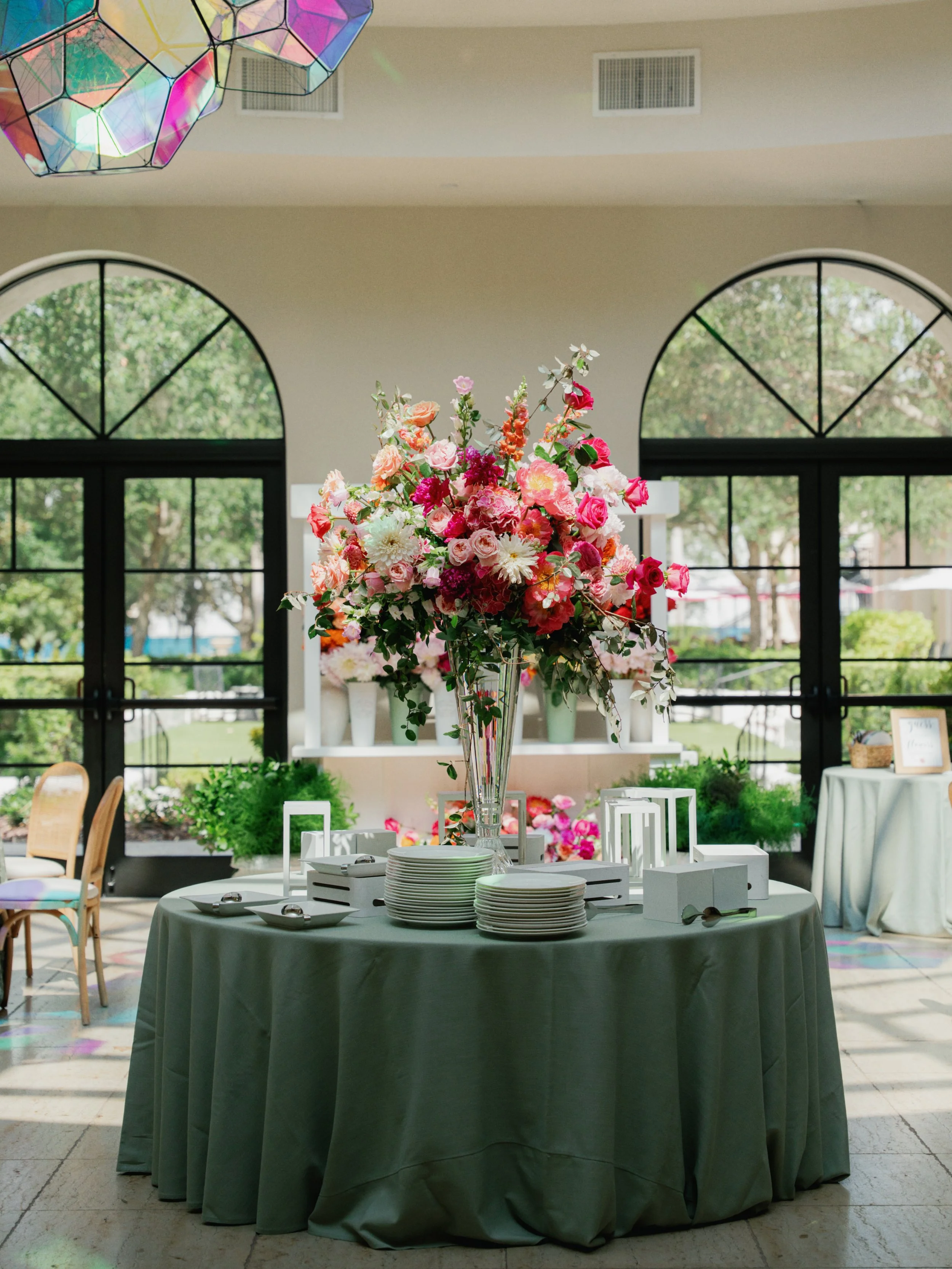 A table with a grey tablecloth featuring a large flower arrangement in a tall clear glass vase, surrounded by white plates, small bowls, and cutlery. The background includes large arched windows with a view of greenery outside and a ceiling with colo