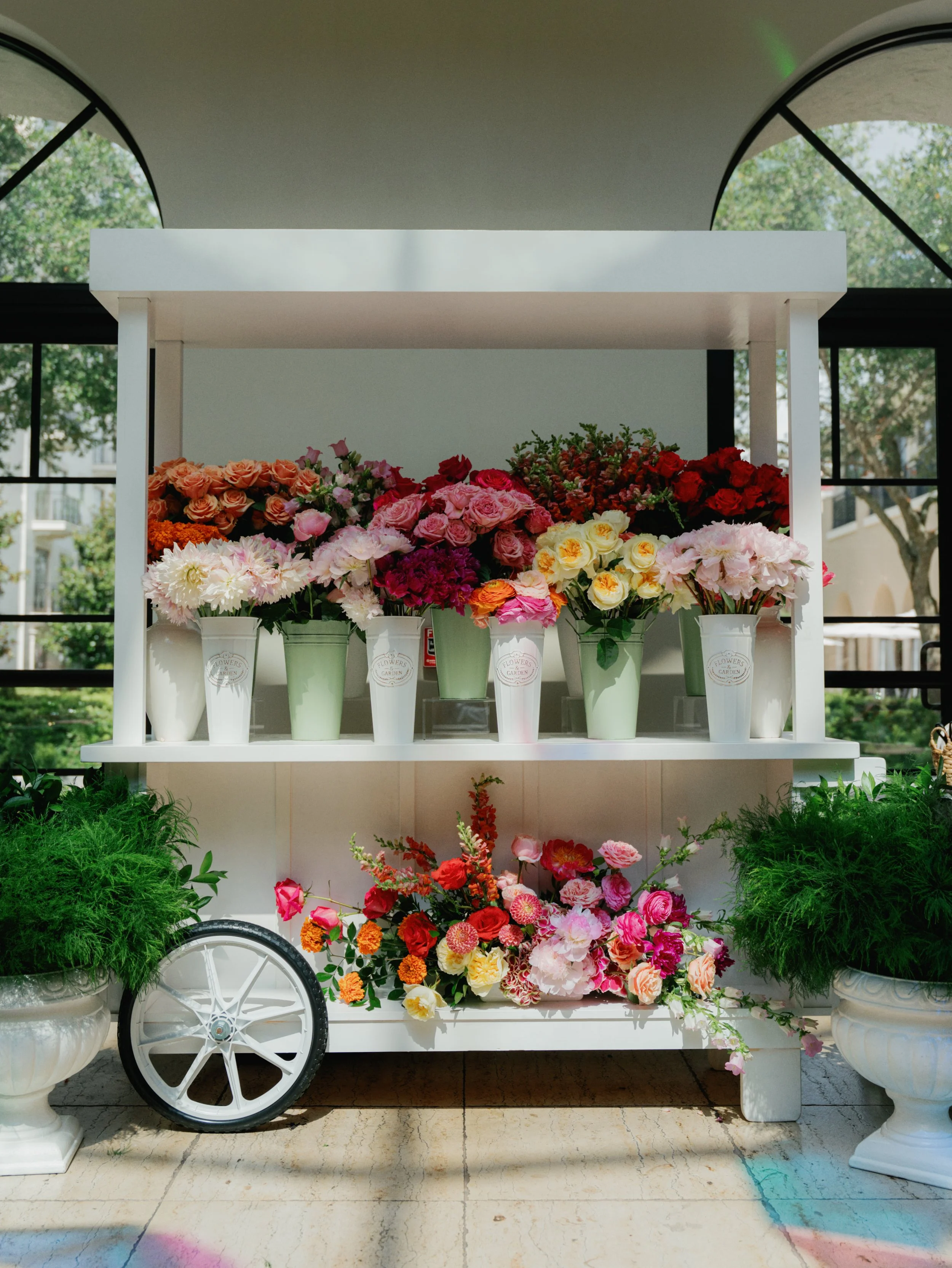 Display of various colorful flowers in white and green vases on a two-tiered white cart, with potted green plants on either side, set outdoors with trees and a building in the background.