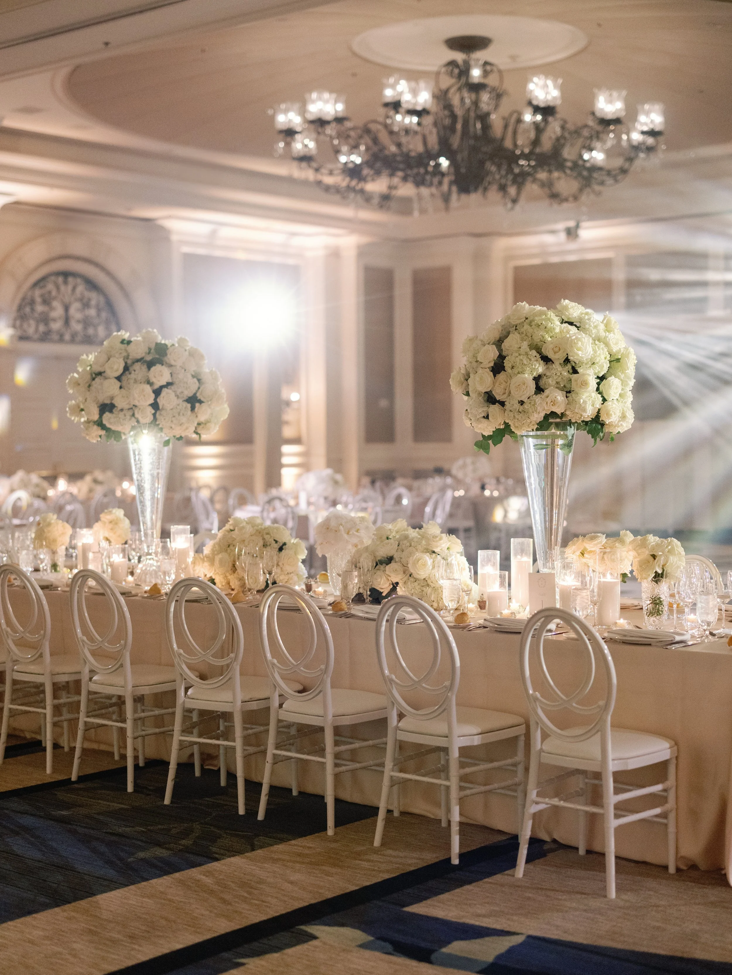Elegant banquet hall decorated for a wedding reception with white floral centerpieces on tall vases, surrounded by candles, and a chandelier hanging from the ceiling.