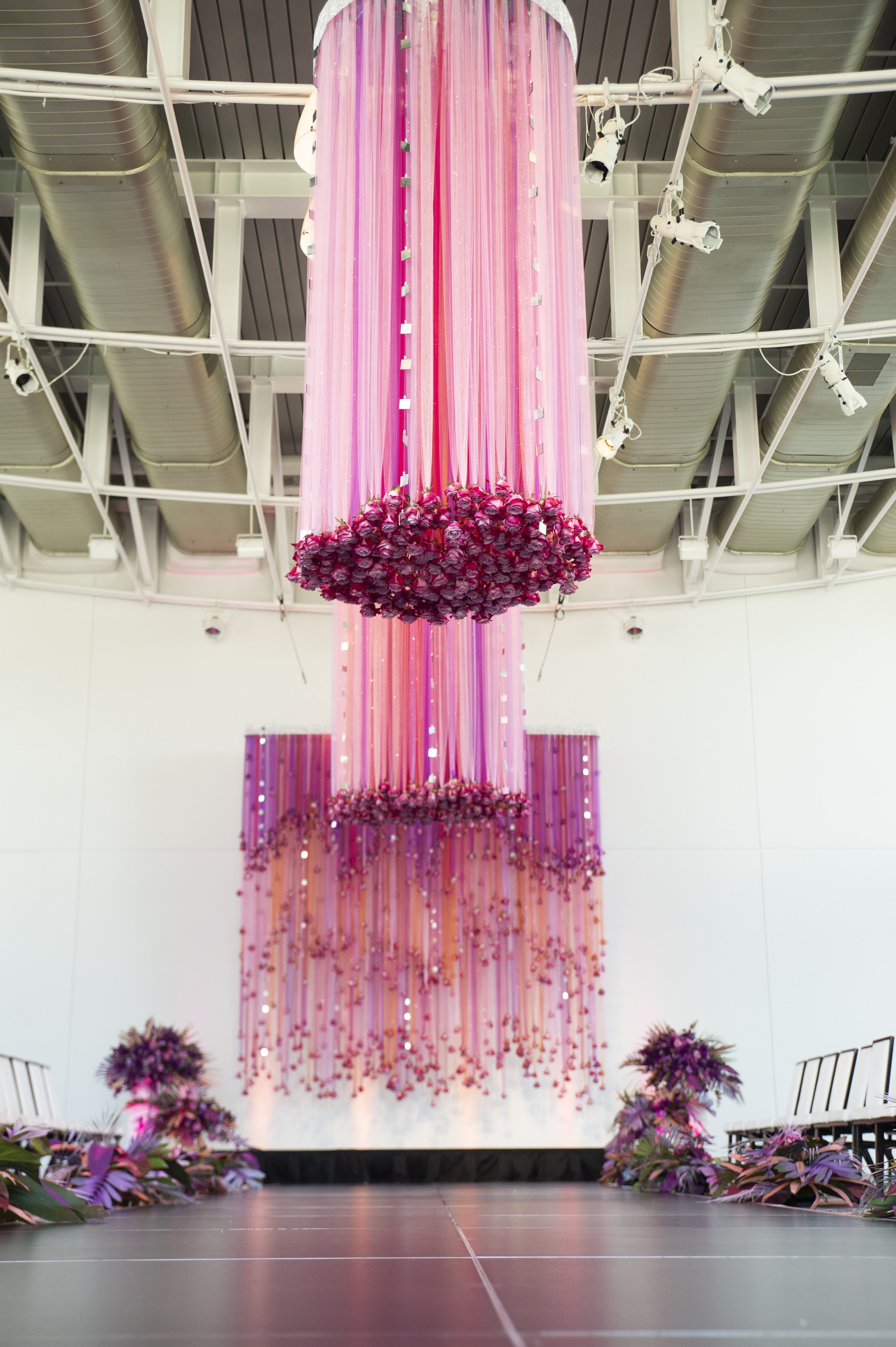 A large pink and purple floral decoration hanging from the ceiling, with a cluster of roses at the bottom center, surrounded by purple flowers and greenery on the stage floor.