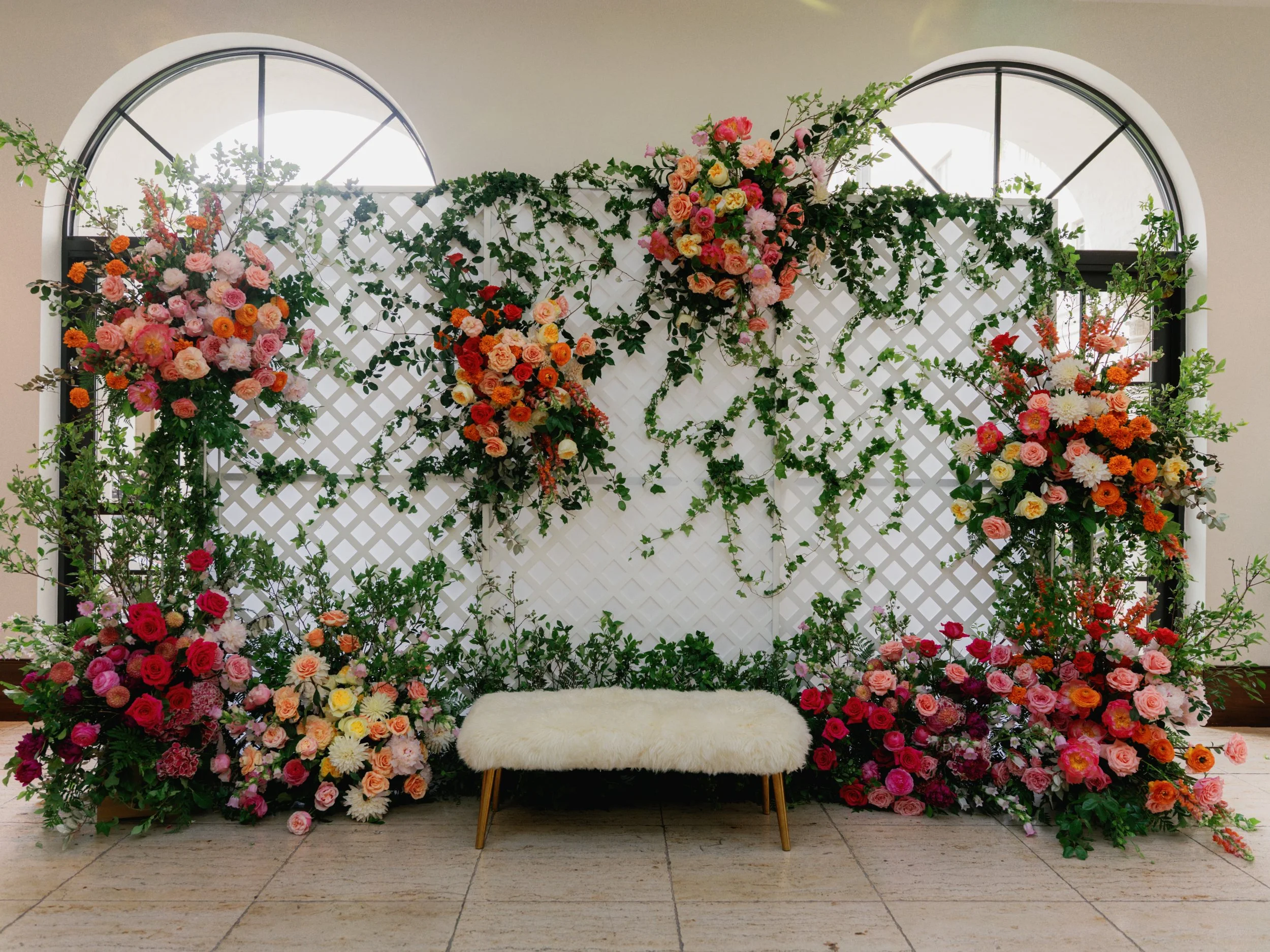 A decorative backdrop with pink, orange, and white flowers and green foliage, featuring large arched windows and a white furry bench in front.