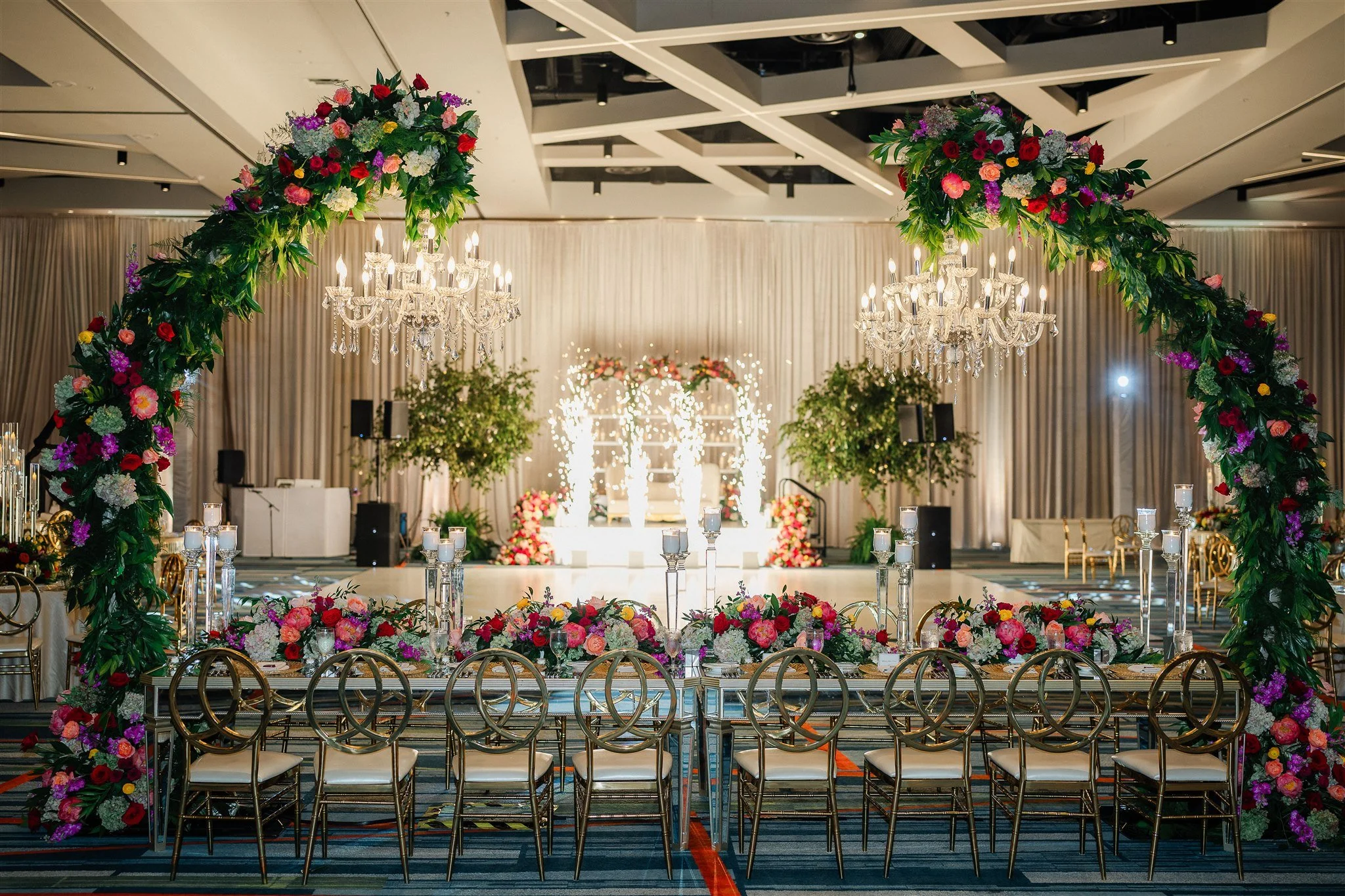 Elegant banquet hall decorated for a wedding or celebration, featuring a floral arch with pink, purple, red, and white flowers, crystal chandeliers, and chairs arranged around long tables with floral centerpieces.