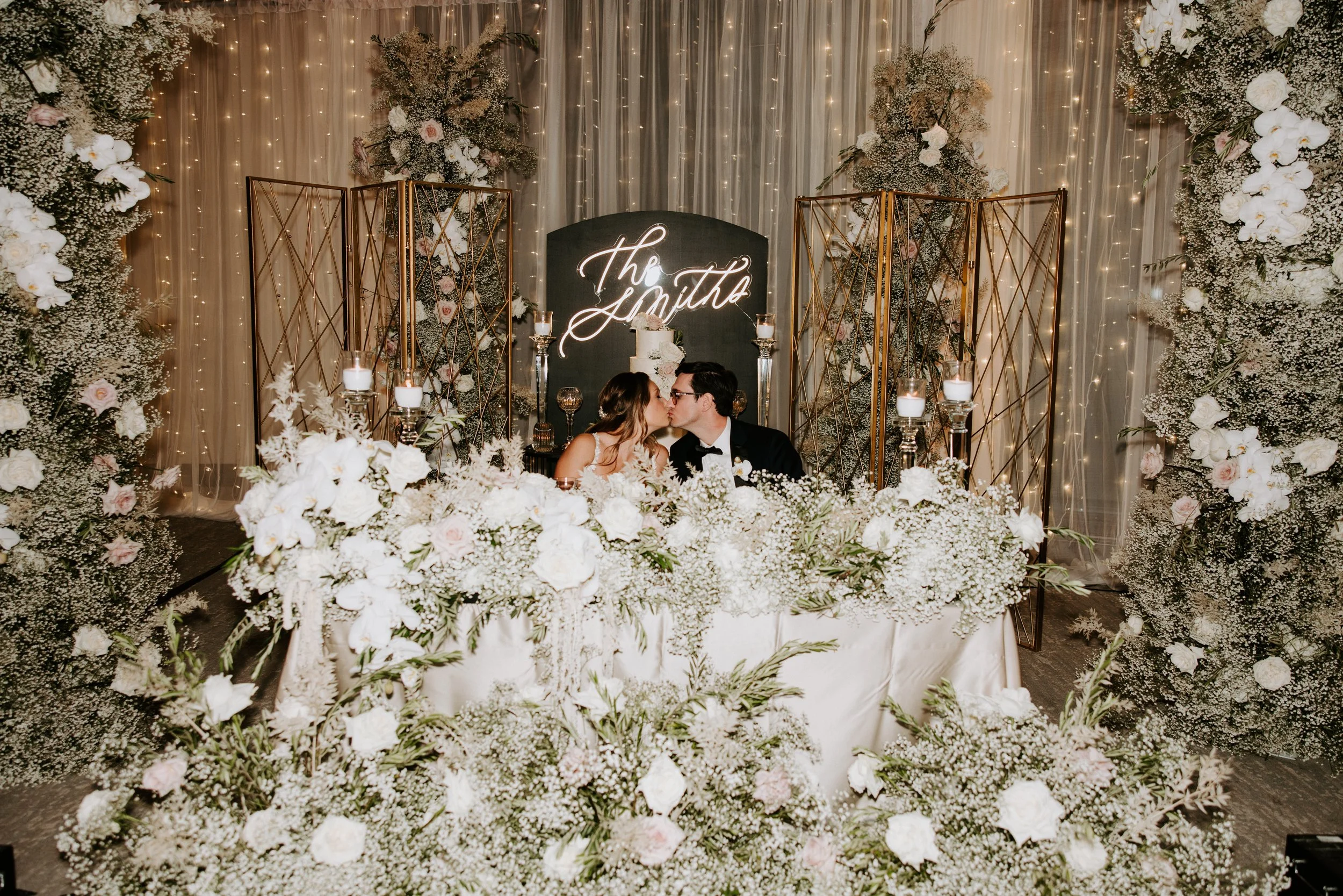 A wedding reception scene with a newly married couple kissing at a table decorated with white flowers. Behind them, a black backdrop with neon signs reading 'The Jones' is surrounded by floral arrangements and warm string lights.