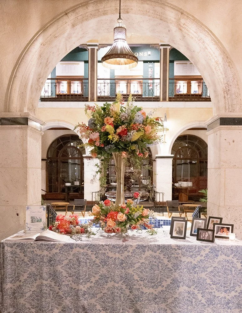 A floral arrangement on a table with framed photographs, an open book, and candles, set in a grand interior with stone arches, a hanging lamp, and a balcony in the background.