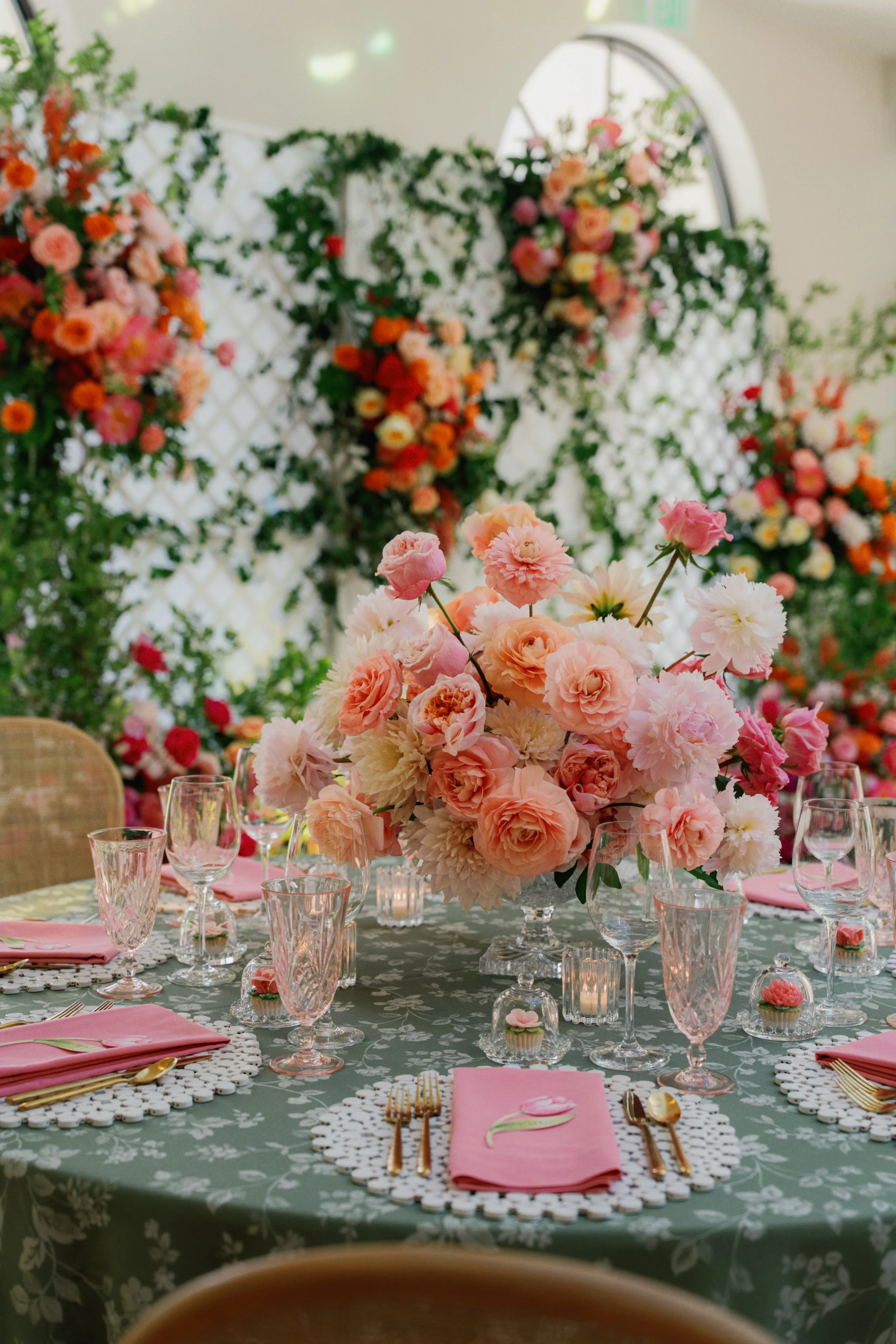 A table decorated for a celebration with a centerpiece of pink and white flowers, pink napkins, gold utensils, and glassware, set on a green tablecloth with a floral pattern, in a room with colorful flowers and greenery in the background.