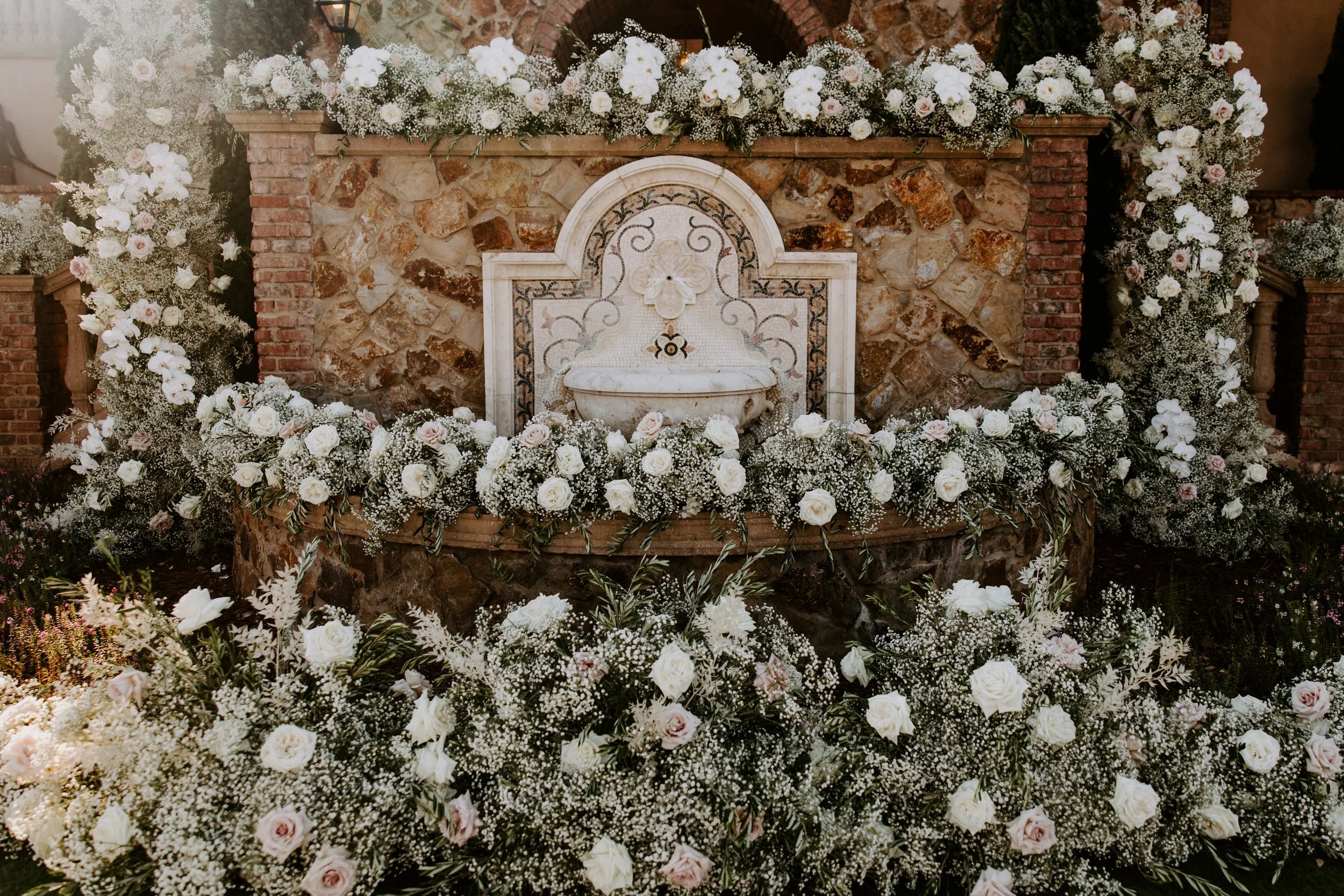A decorative stone and brick wall with a fountain or basin in the center, surrounded by an abundance of white and pale pink flowers, likely for a wedding or special event.