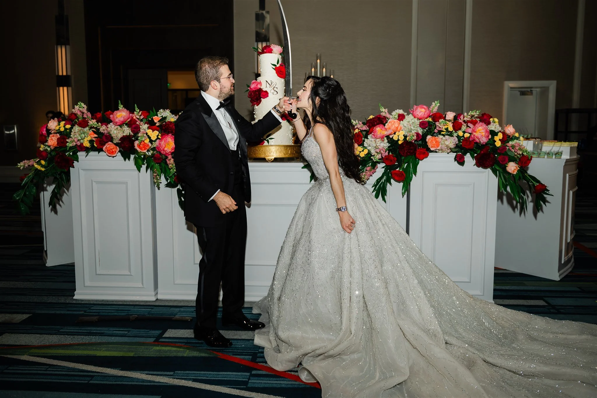 A bride and groom sharing a moment with a wedding cake in the background, decorated with flowers, at their wedding reception.