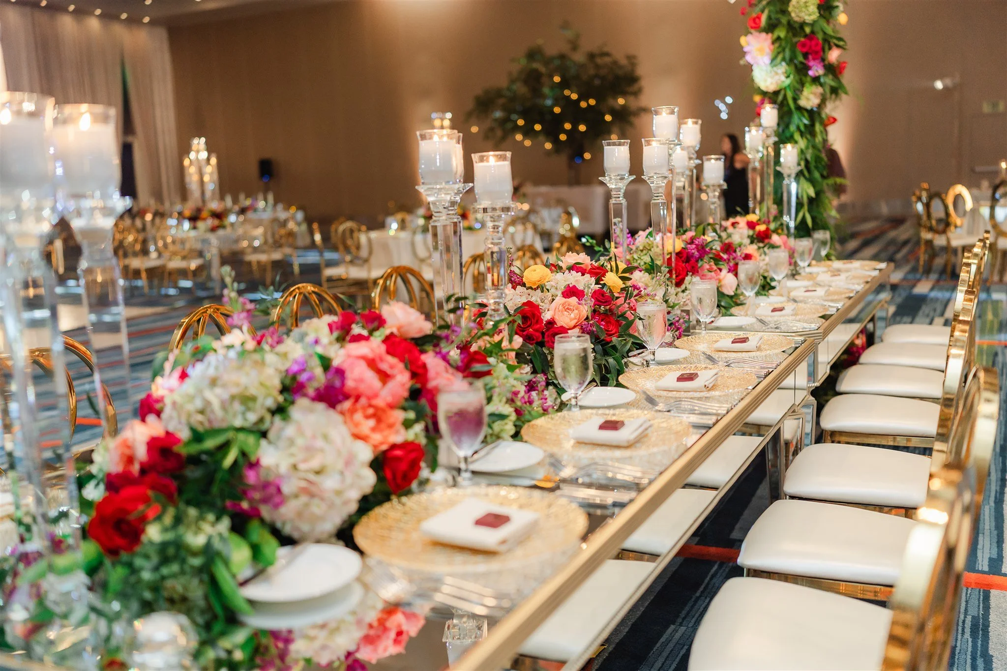 Long banquet table decorated with colorful flowers, tall candleholders with lit candles, and place settings with white plates, gold chargers, and napkins. The background shows a softly lit room with blurred figures and a festive atmosphere.