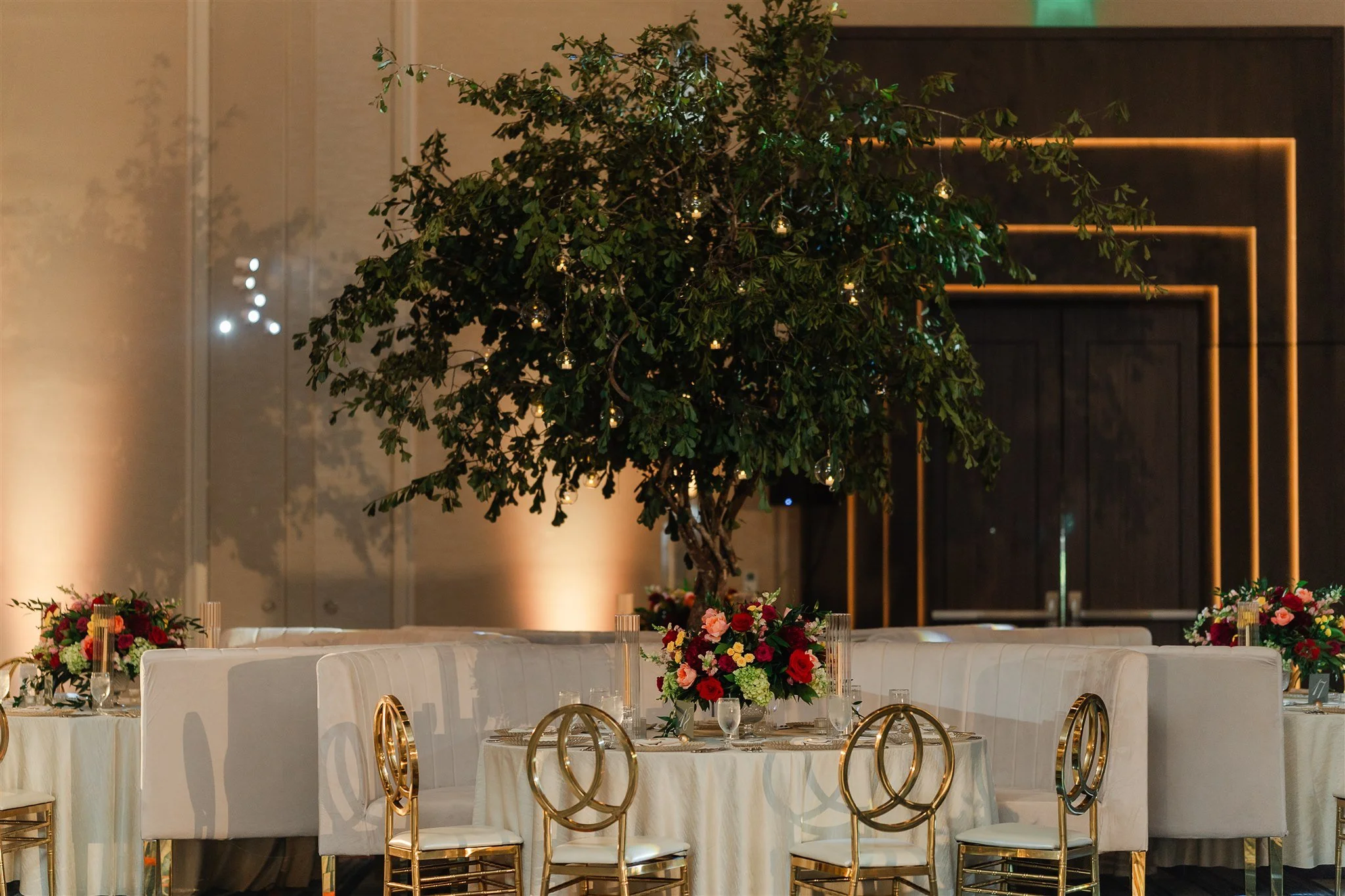 Decorated event space with a large indoor tree adorned with small hanging lights, surrounded by tables with floral centerpieces, gold chairs, and elegant tableware, in a modern venue with dim lighting.