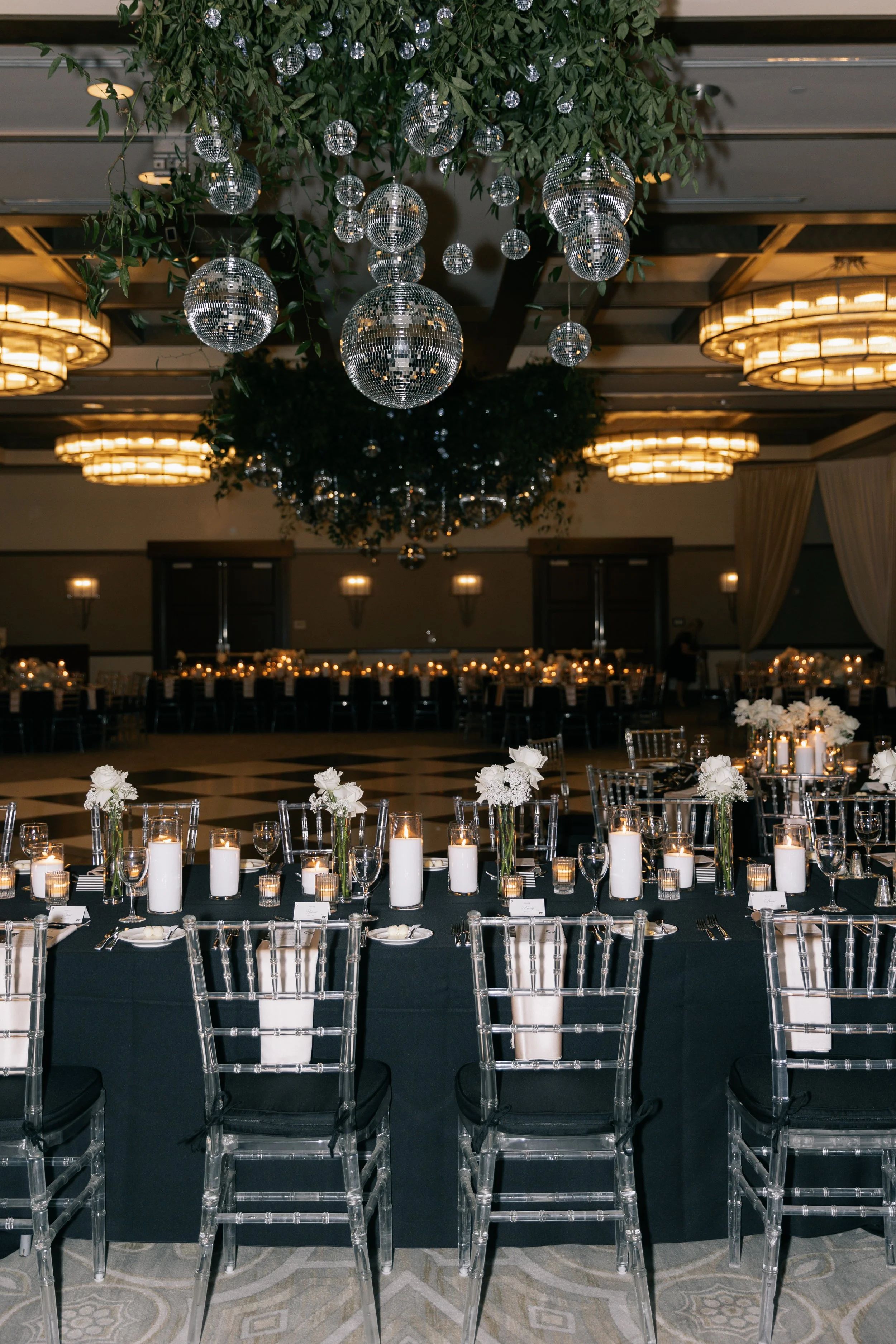 Elegant banquet hall decorated with white flowers, candles, and glass chairs, featuring a ceiling installation of disco ball ornaments and warm lighting.