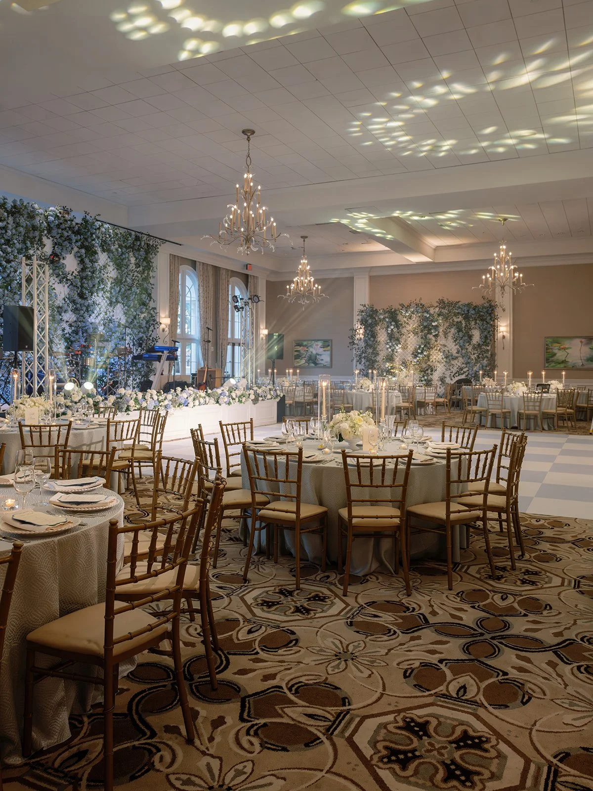 Elegant banquet hall decorated for a wedding or event with round tables covered in white tablecloths, surrounded by wooden chairs with cushions, adorned with floral centerpieces, candles, and lit chandeliers reflecting light onto the ceiling. The bac
