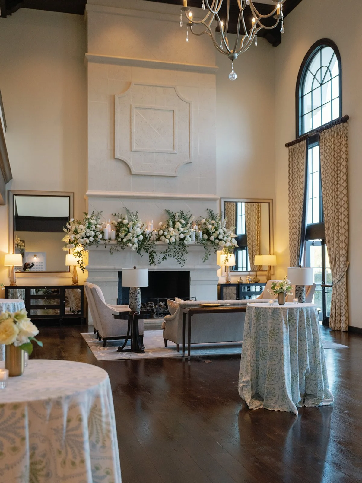 Elegant living room with large arched windows, a white fireplace decorated with flower arrangements, candles, and gold accents, surrounded by comfortable furniture and decorative lamps.