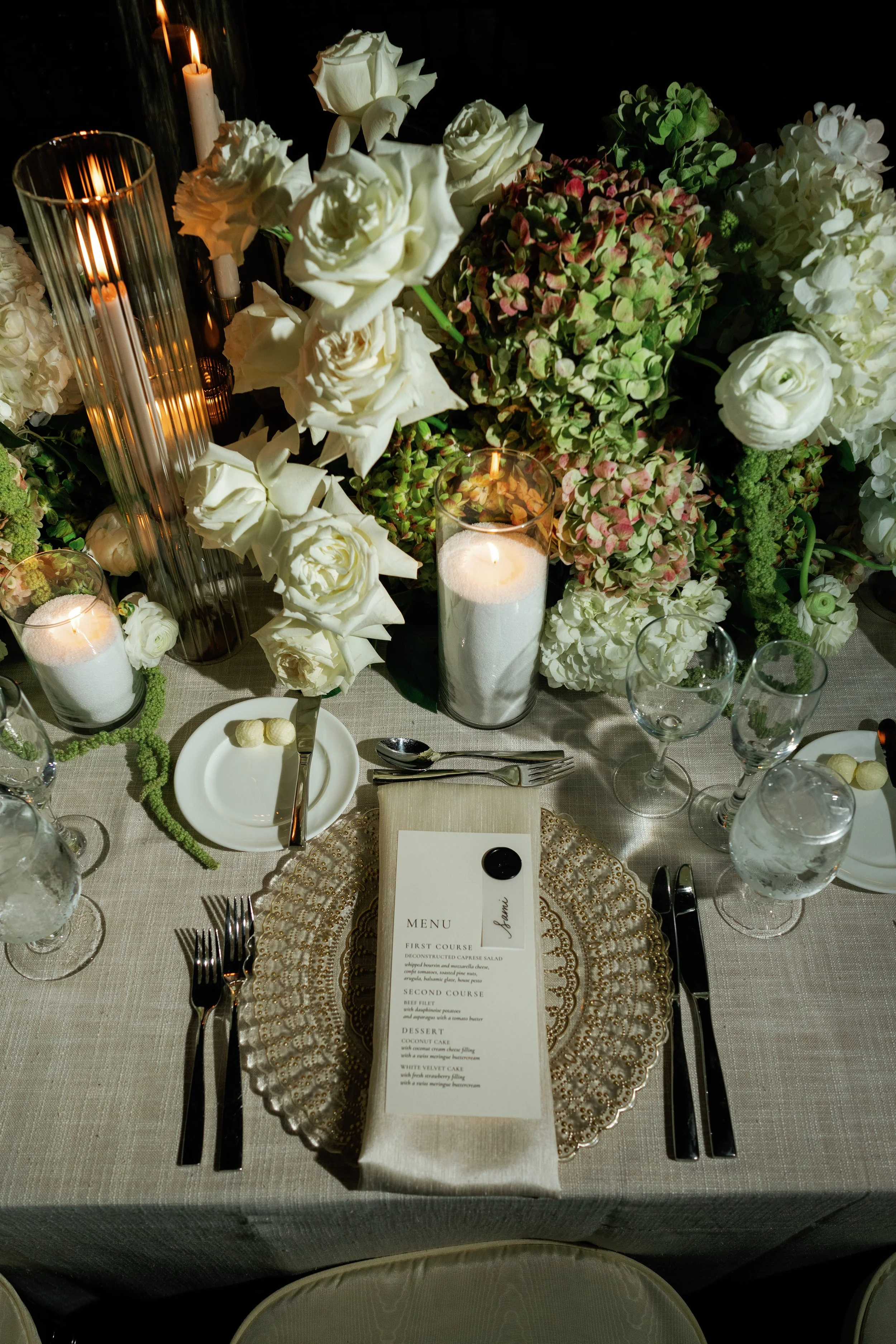 A formal dining table setup with a menu card placed on a decorative gold charger plate, surrounded by crystal wine glasses, cutlery, and white candles. The table is decorated with various white and green floral arrangements, including roses, hydrange